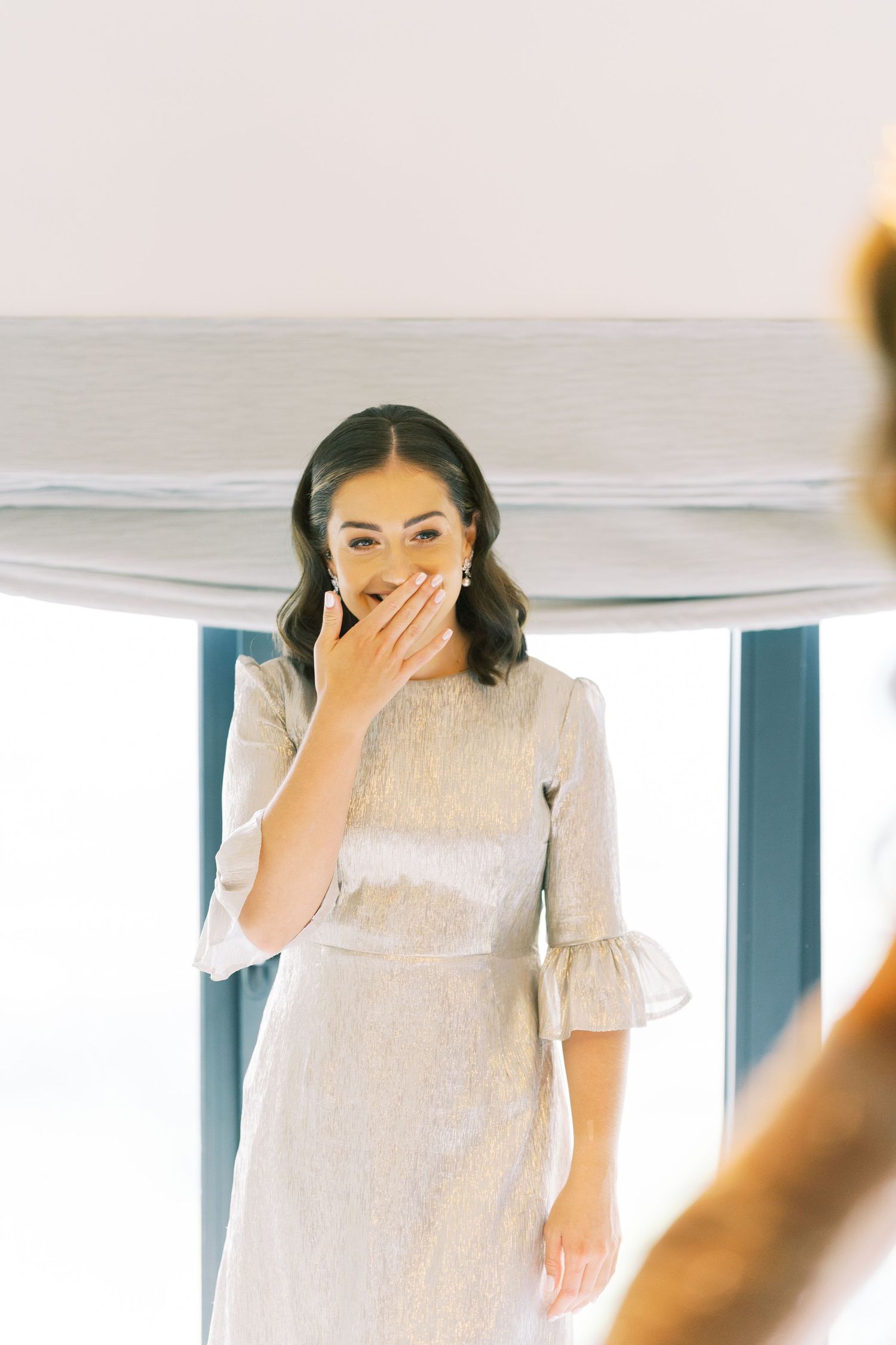 Bride in silver dress covers mouth in surprise during wedding preparations.