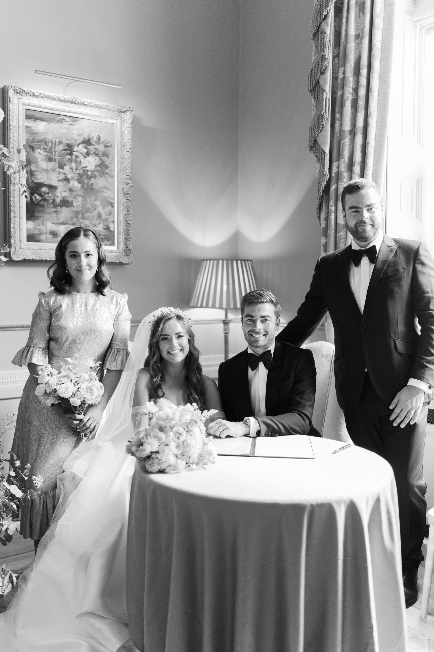 Bride and groom signing wedding register at table, flanked by bridesmaid and best man in elegant venue.
