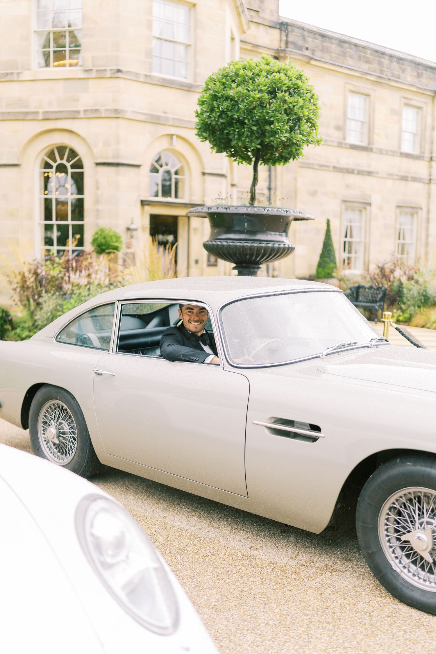 A person drives a classic silver Aston Martin DB5 outside an elegant stone manor house with topiary gardens.