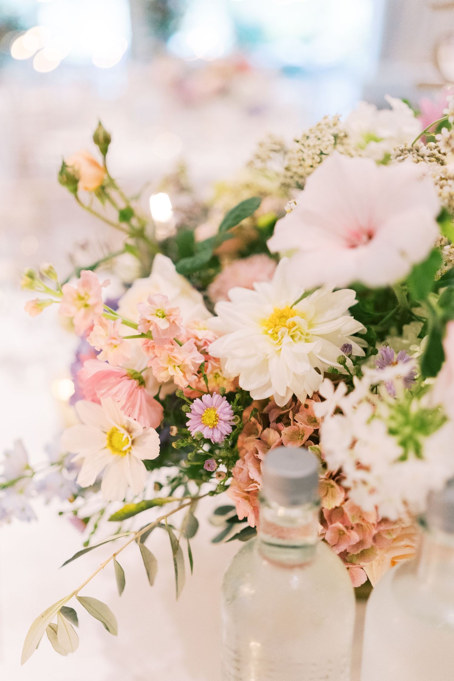 Soft pastel wedding floral centerpiece with white daisies, pink blooms, and greenery on a reception table.