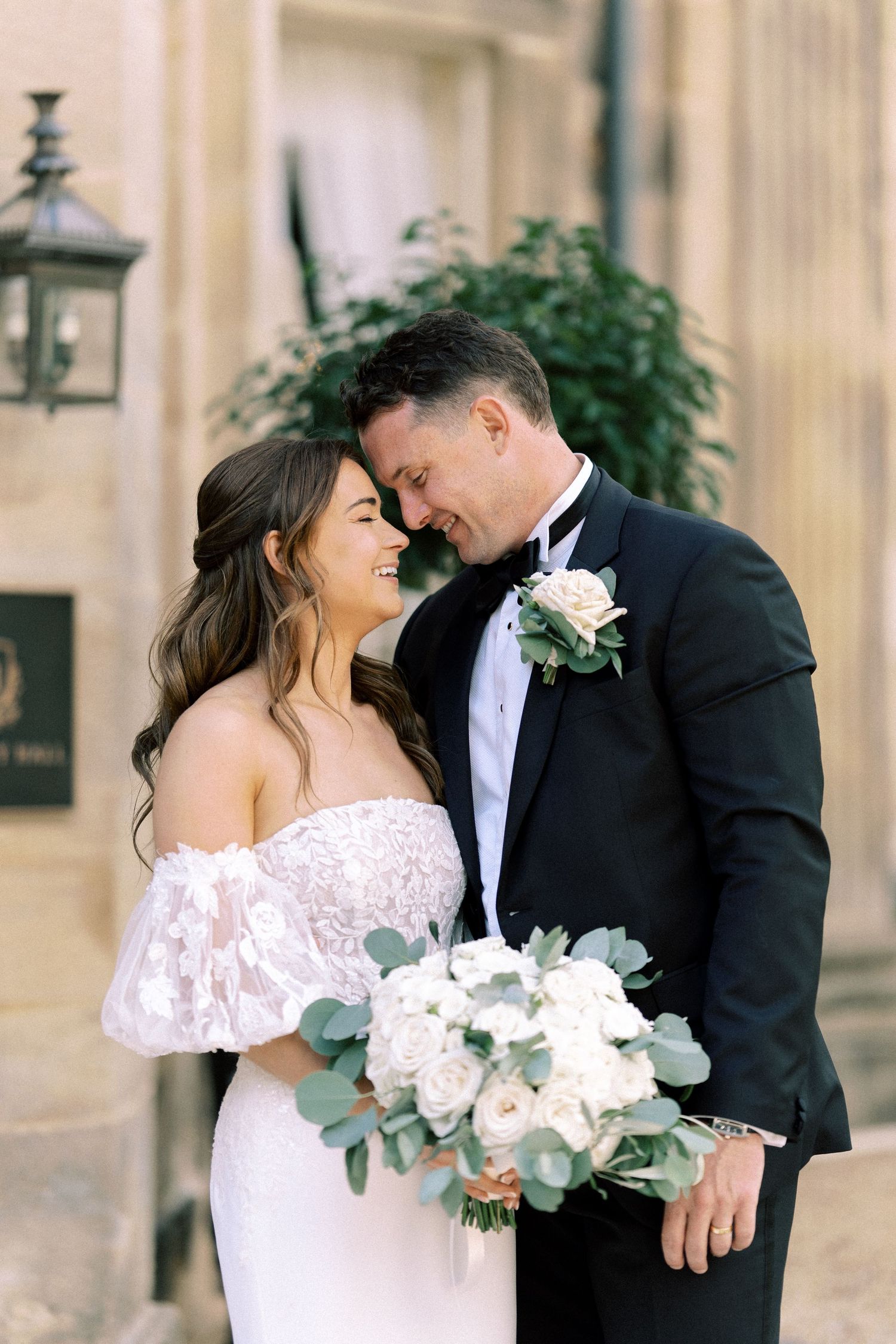 Smiling bride and groom share a tender moment, bride holding white rose and eucalyptus bouquet.