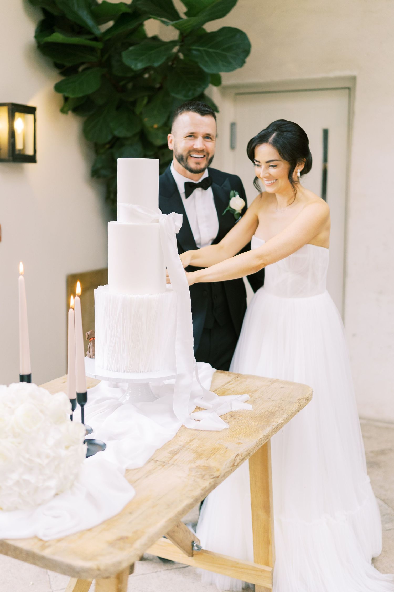 Happy couple cutting white tiered wedding cake at candlelit table with greenery backdrop.