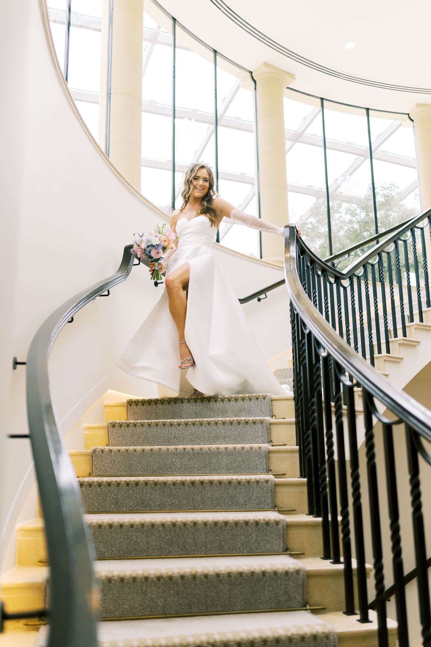 Bride in white gown holding bouquet descends elegant curved staircase with black iron railing and large windows.