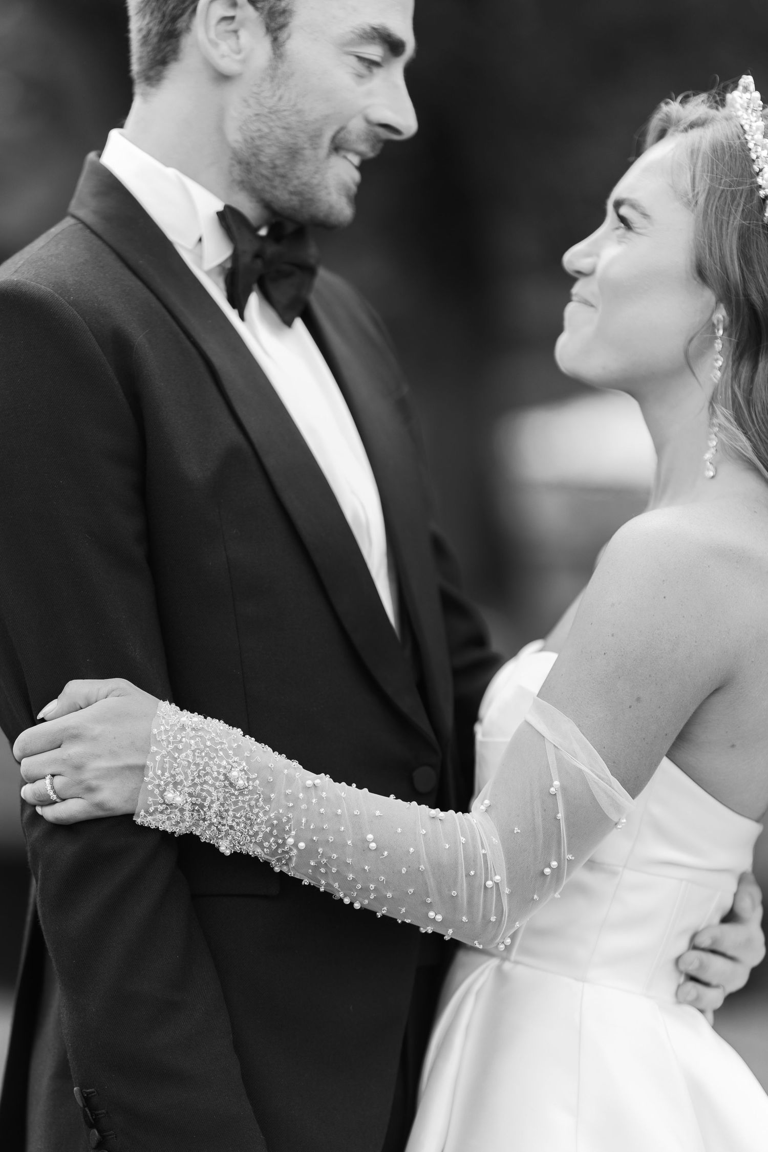 Bride in pearl-embellished gloves and groom in black tuxedo smiling at each other in a black and white portrait.