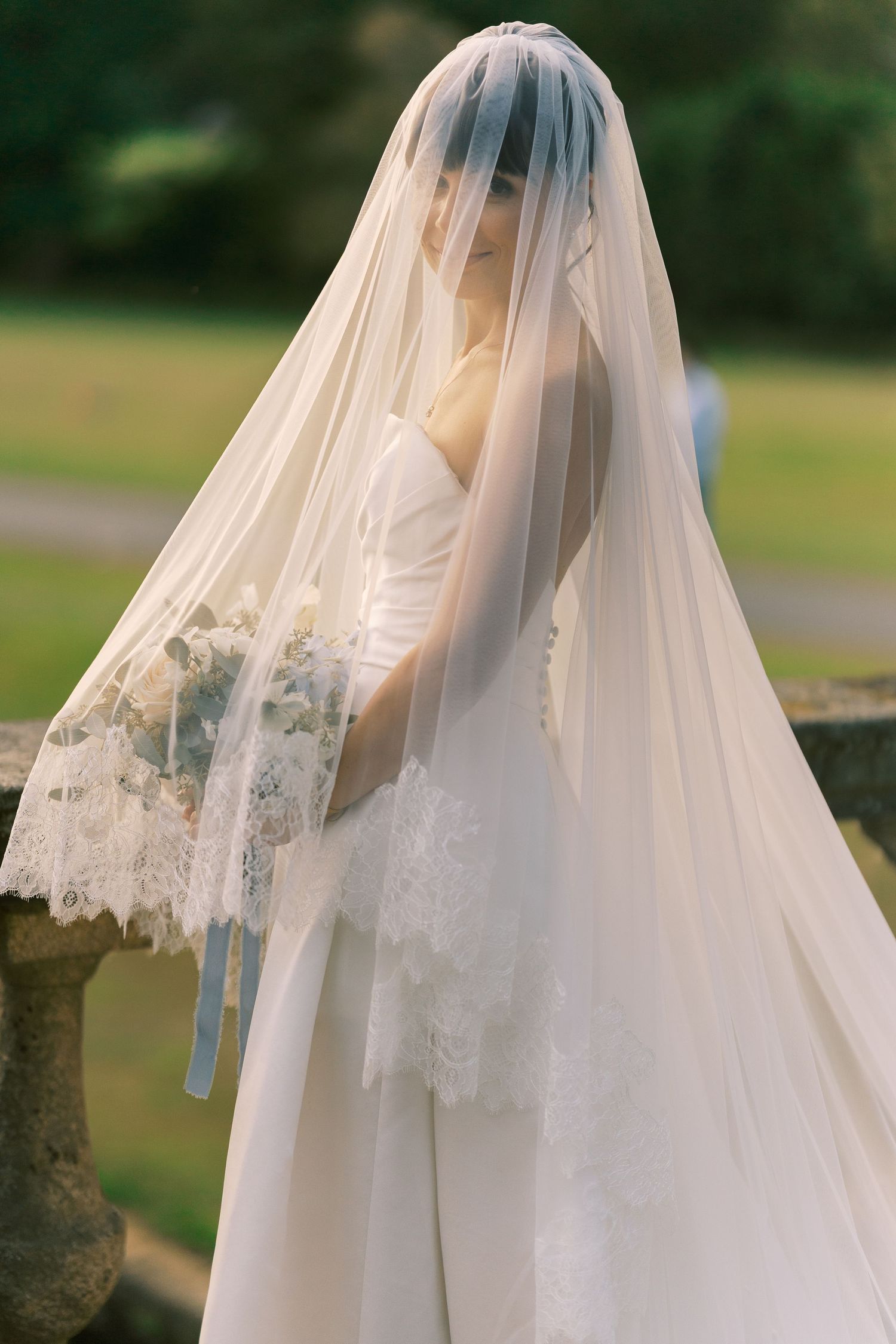 Bride in white lace wedding dress holding bouquet, face covered by long veil with lace trim, outdoors.
