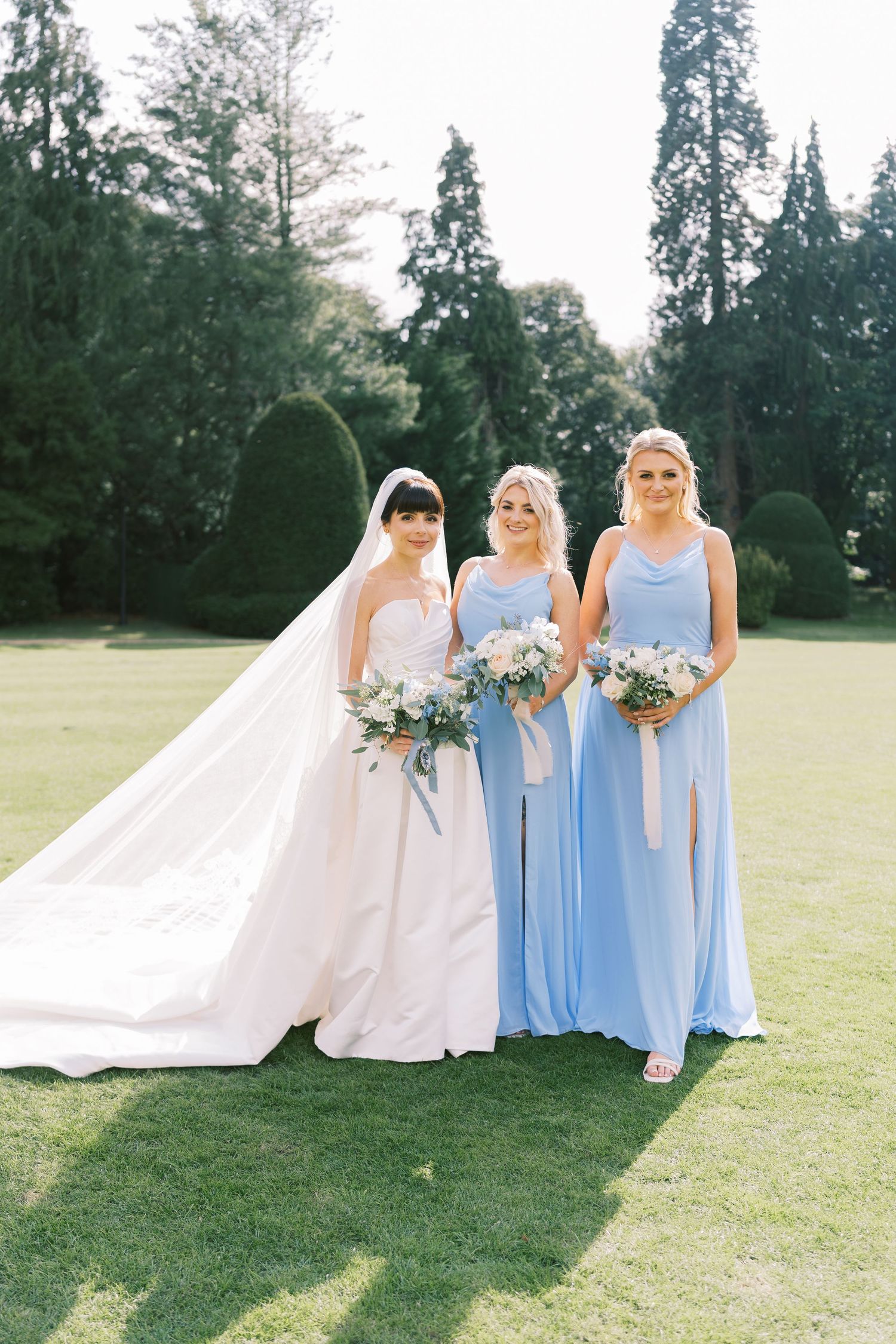 Bride in white gown with veil poses with two bridesmaids in light blue dresses on green lawn.