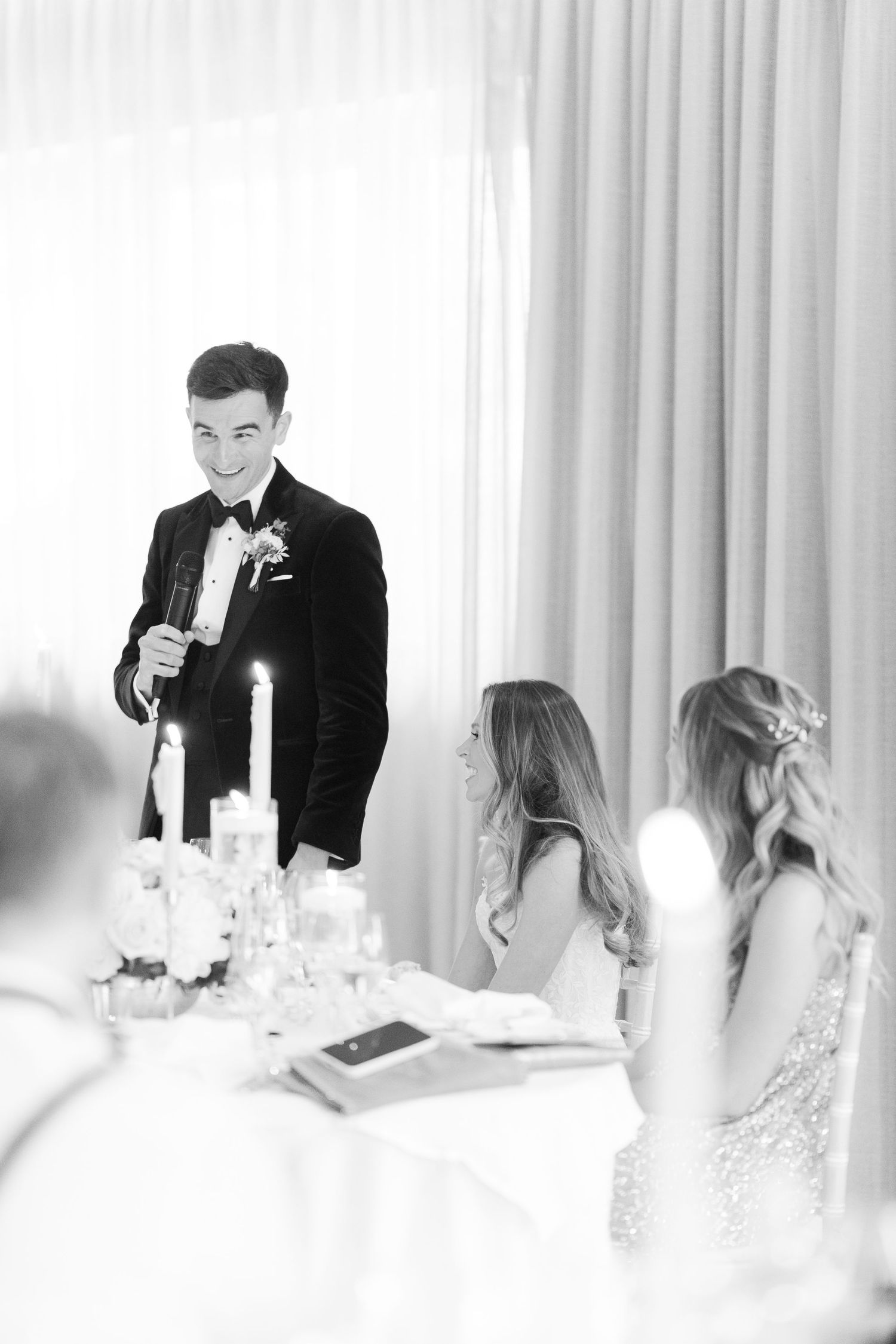 Groom in black tuxedo gives speech at candlelit wedding reception table in black and white photo.