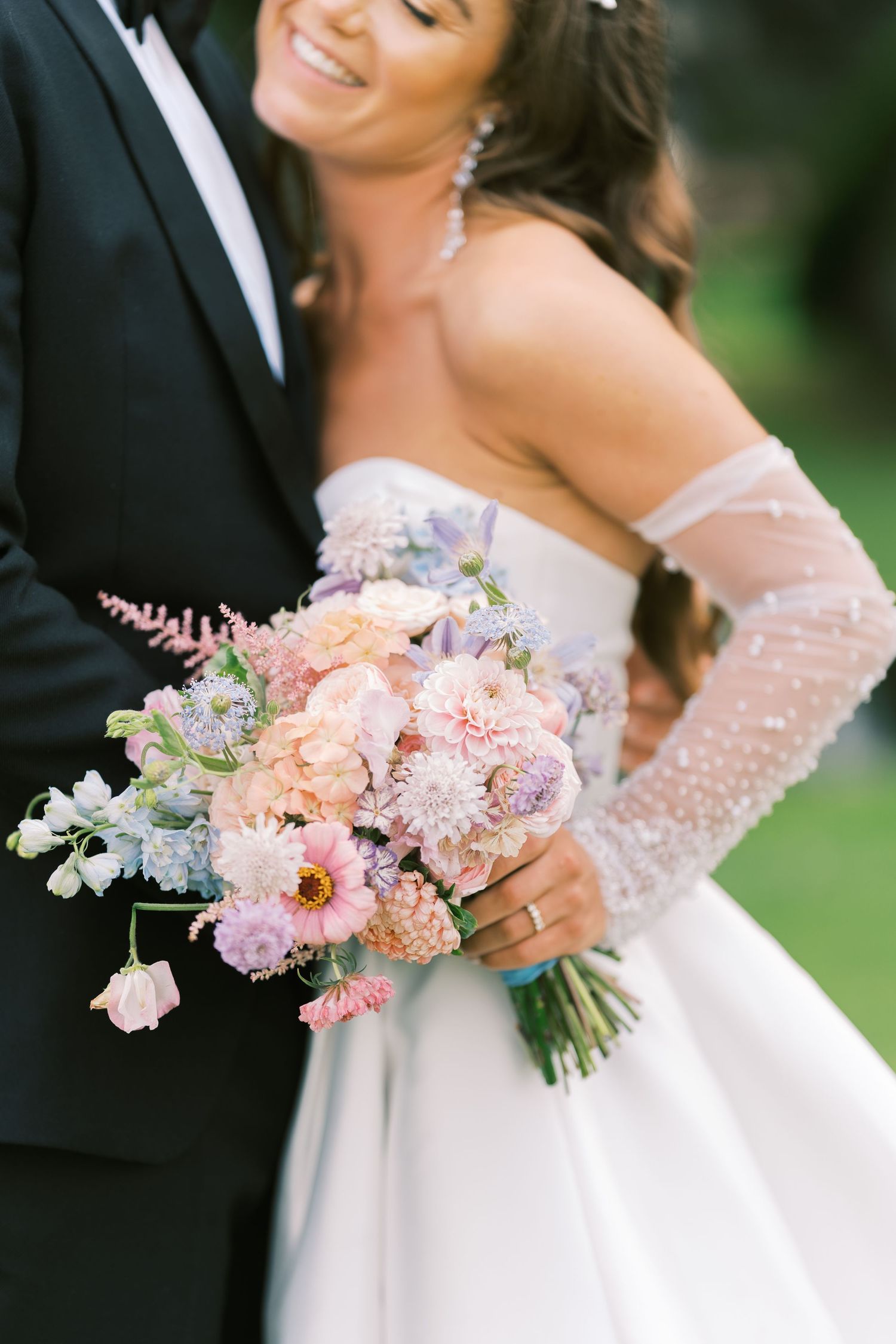 Close-up of bride holding lush pastel bouquet of pink, lavender, and peach blooms in beaded off-shoulder gown.