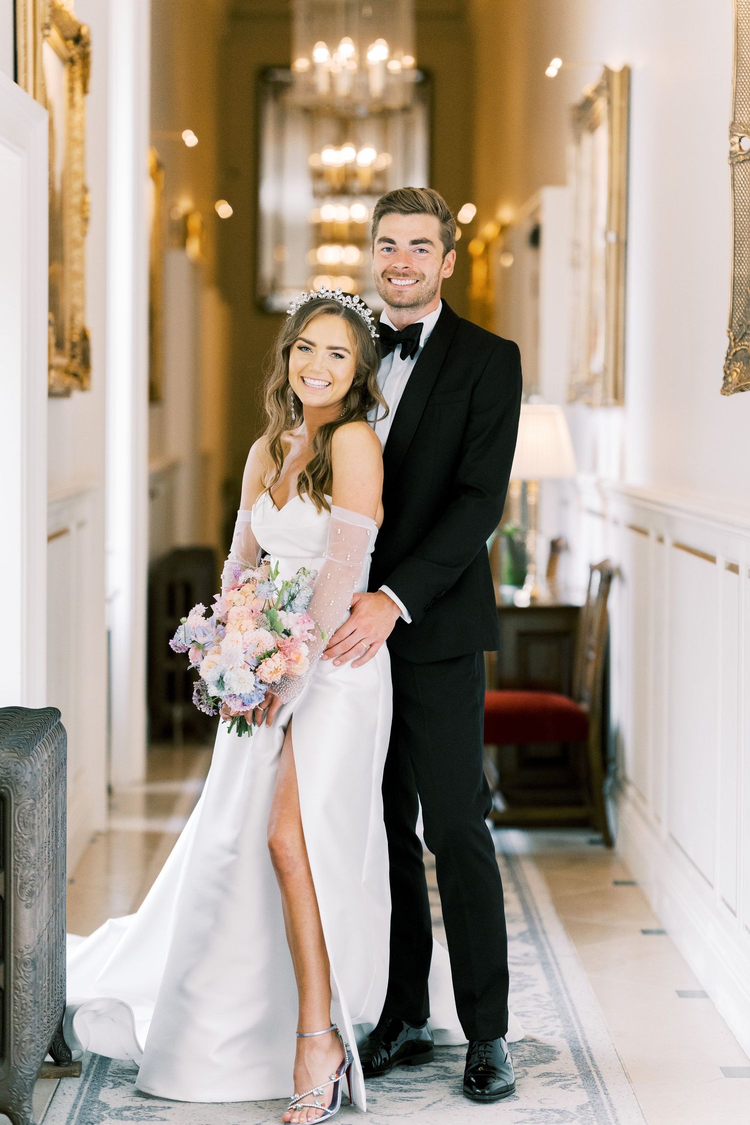Bride and groom posing in ornate hallway, she holds pink bouquet wearing slit gown, he wears tuxedo.