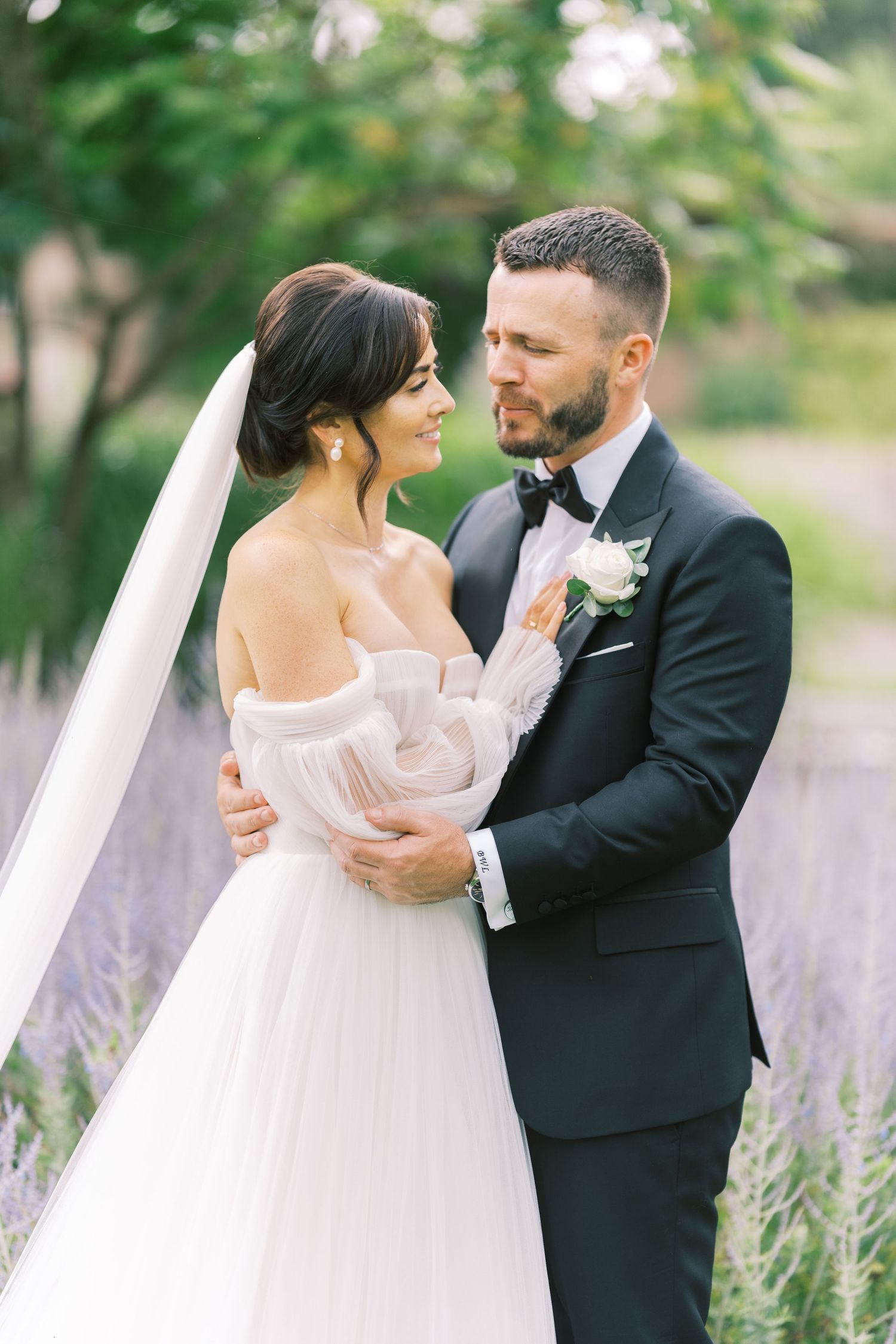 A bride and groom embrace lovingly outdoors, she in an off-shoulder tulle gown with veil, he in a black tuxedo with boutonniere.