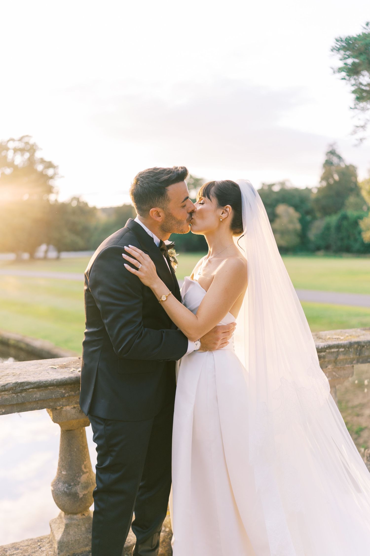 A bride and groom share a romantic kiss at golden hour, with the bride wearing a white gown and veil outdoors.