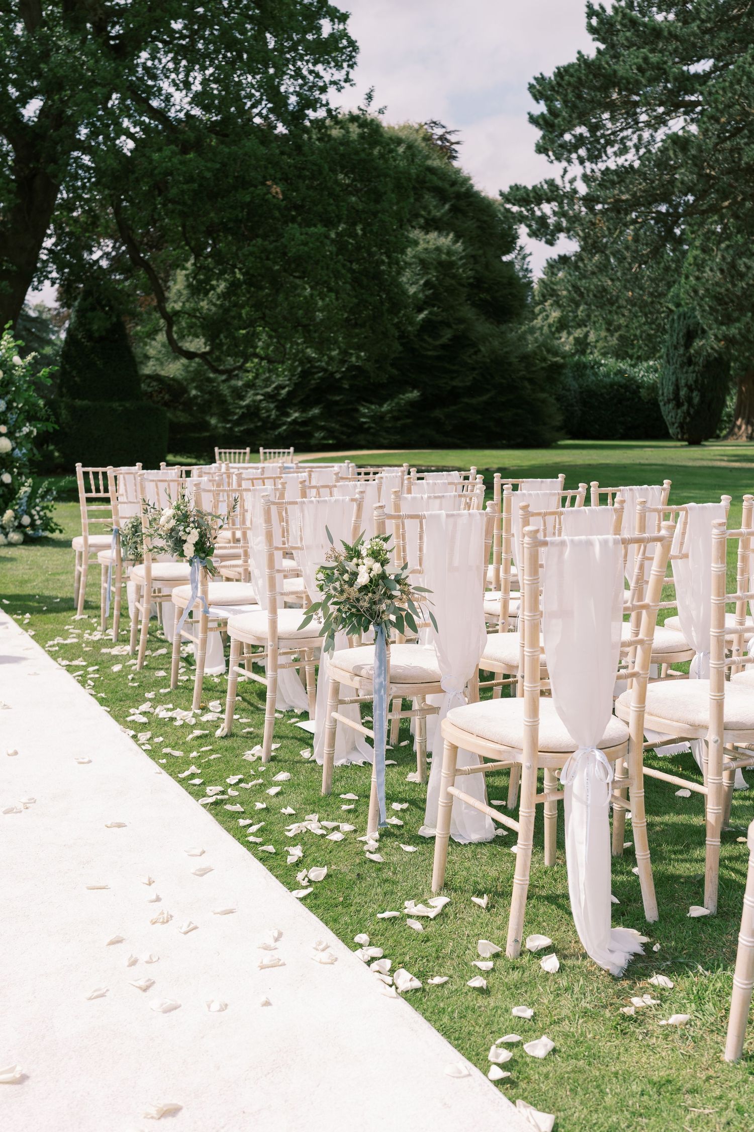 Rows of white chiavari chairs with floral decorations line petal-strewn aisle at outdoor garden wedding.