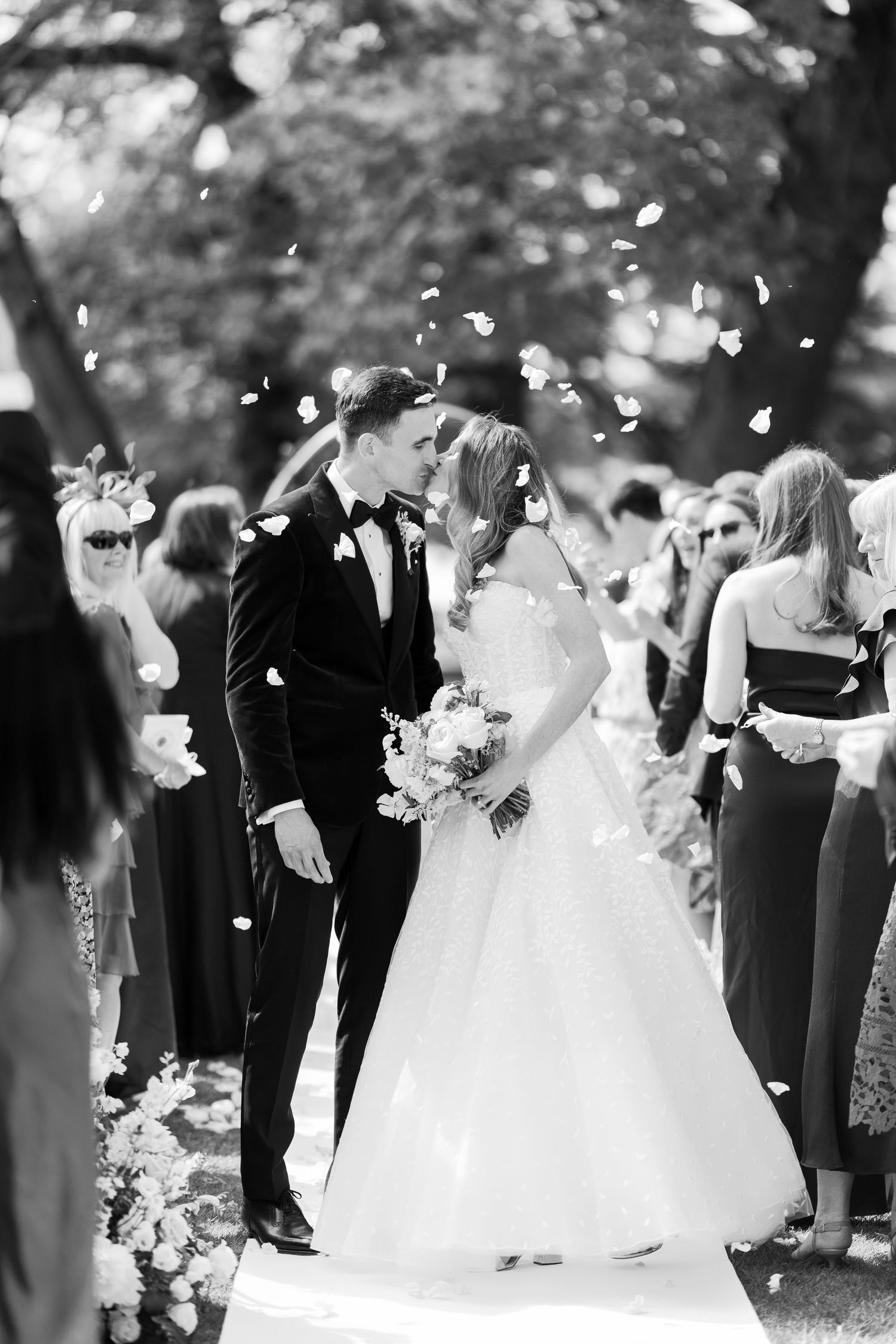 Newlyweds share a kiss as confetti falls during their outdoor wedding ceremony recessional in black and white.