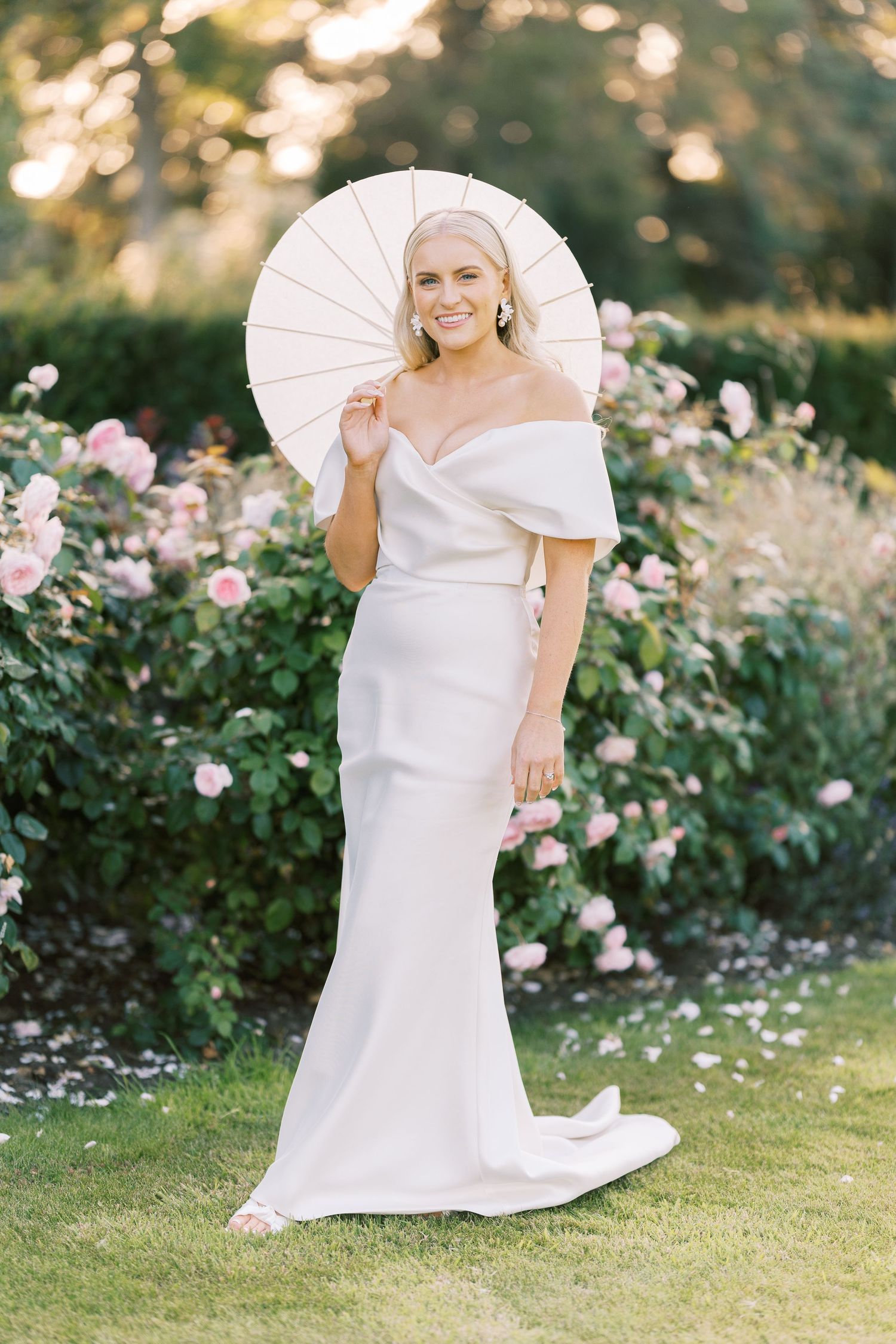 Elegant bride in off-shoulder gown holding white parasol among blooming pink roses at sunset.