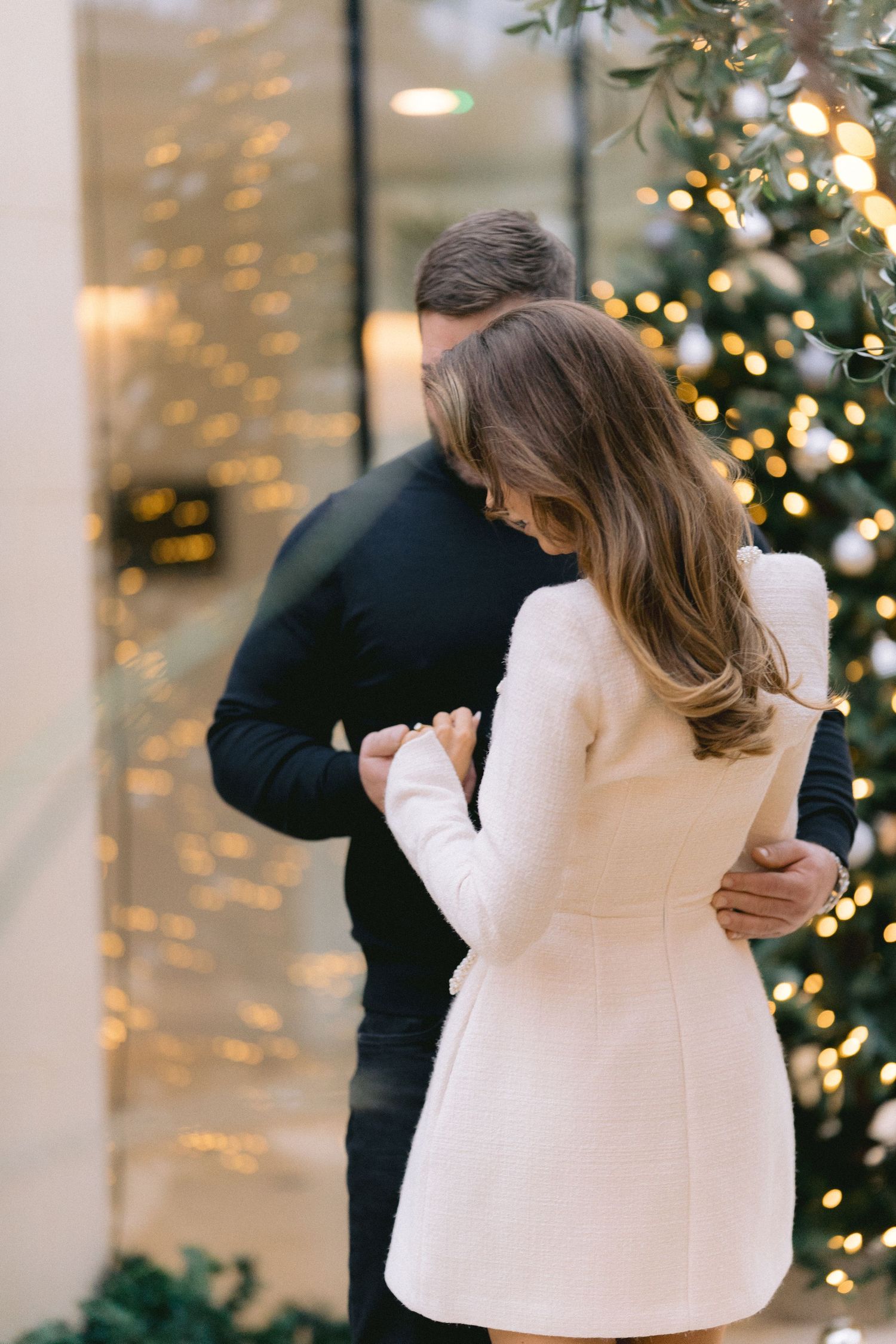 Romantic couple embracing near a decorated Christmas tree with warm bokeh lights in an elegant holiday setting.