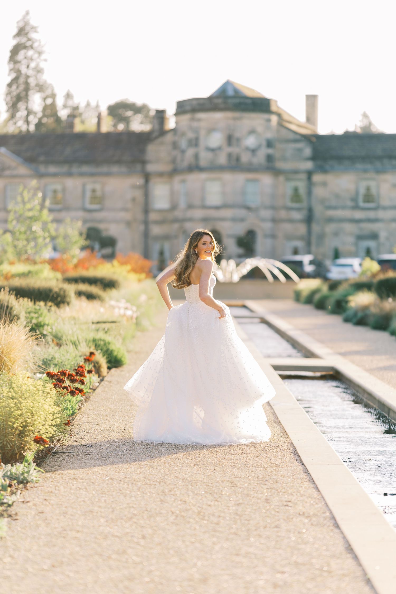 Bride twirls in a stunning layered white ball gown in a sunlit formal garden outside an elegant estate.