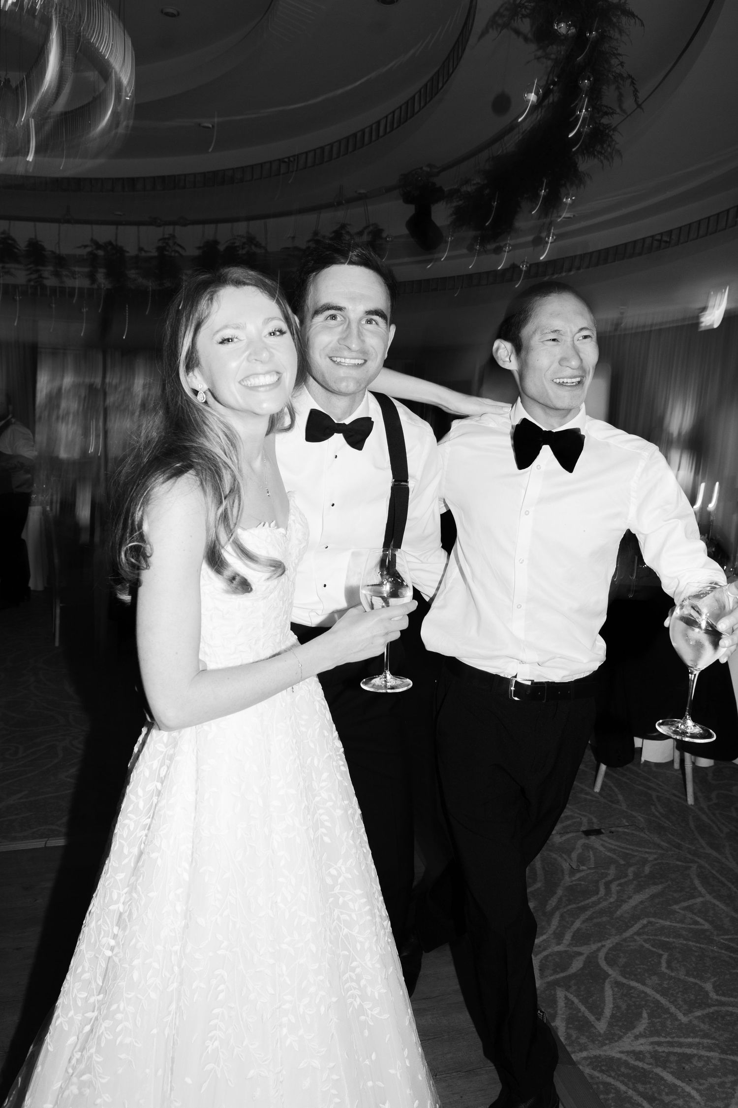 Smiling bride poses with two tuxedo-clad groomsmen holding champagne glasses at wedding reception.