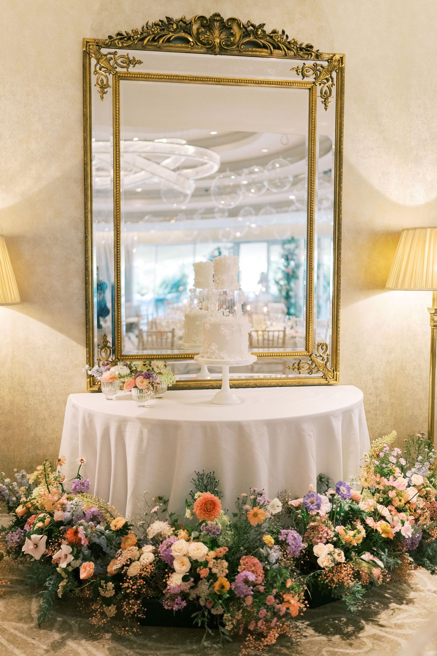 Elegant wedding cake on draped table with colorful floral arrangements and ornate gold mirror backdrop in reception hall.
