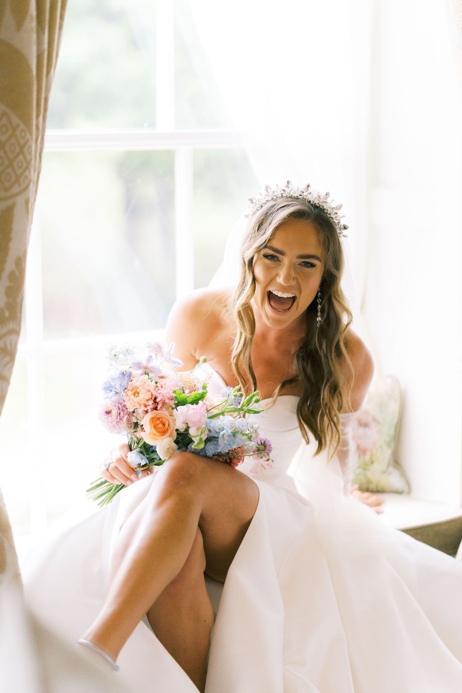 Joyful bride in white gown and tiara laughing while holding colorful pastel bouquet seated by bright window.