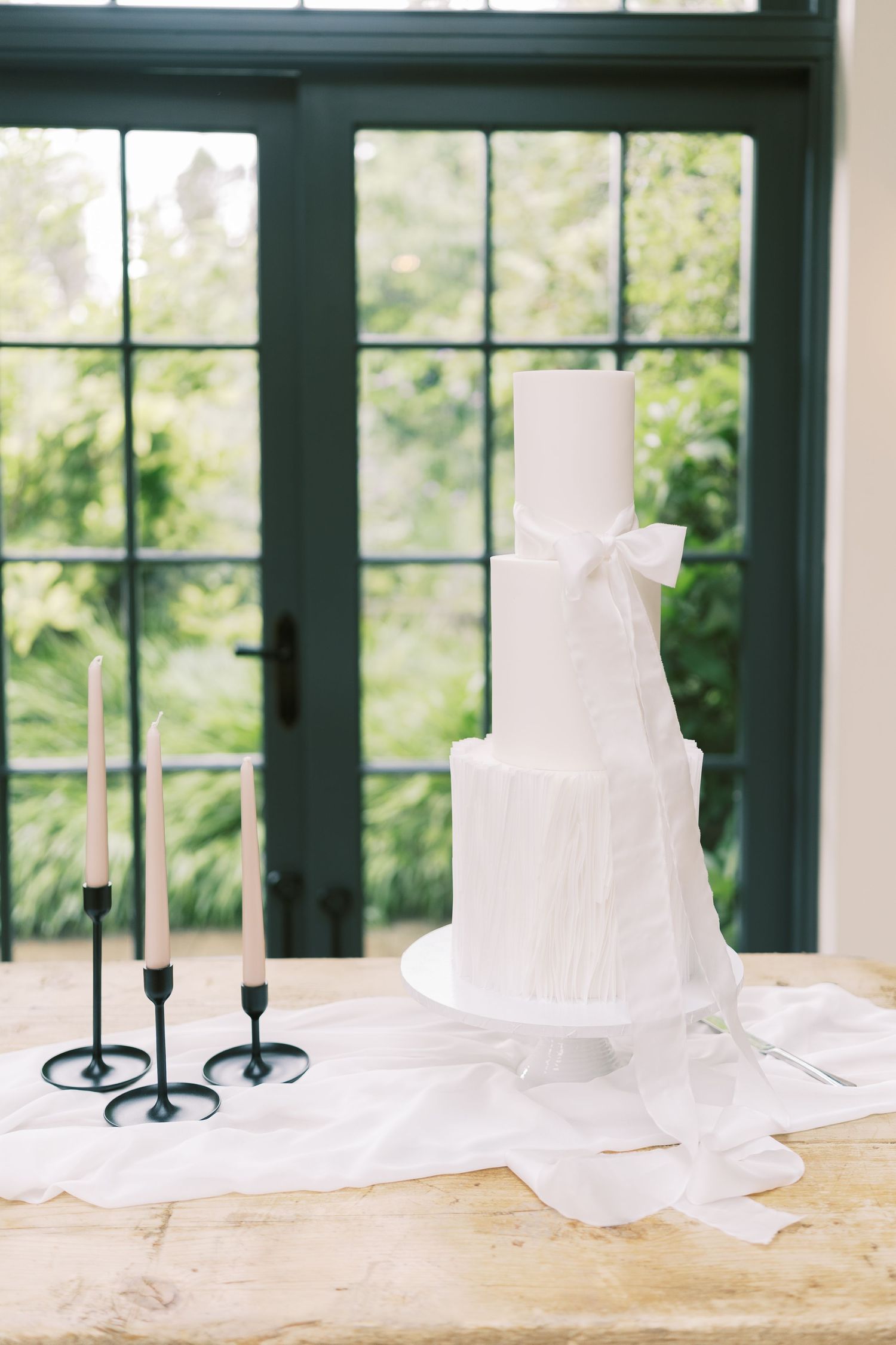 Elegant three-tier white wedding cake with bow, beside black taper candle holders on a rustic wood table.