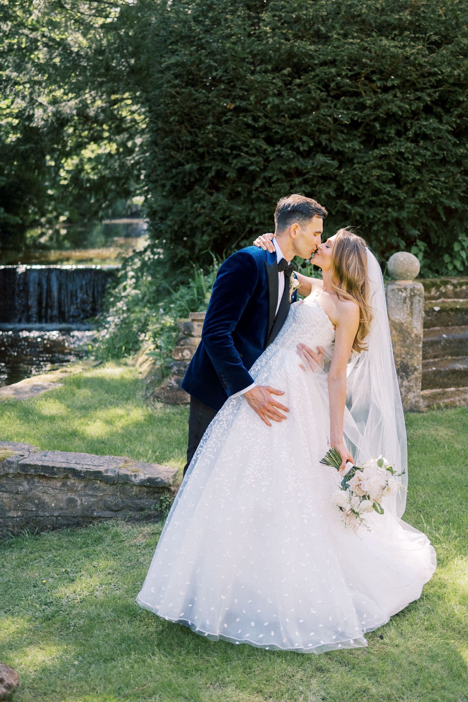 A bride and groom share a romantic kiss in a lush garden, she wears a ballgown and holds a bouquet.