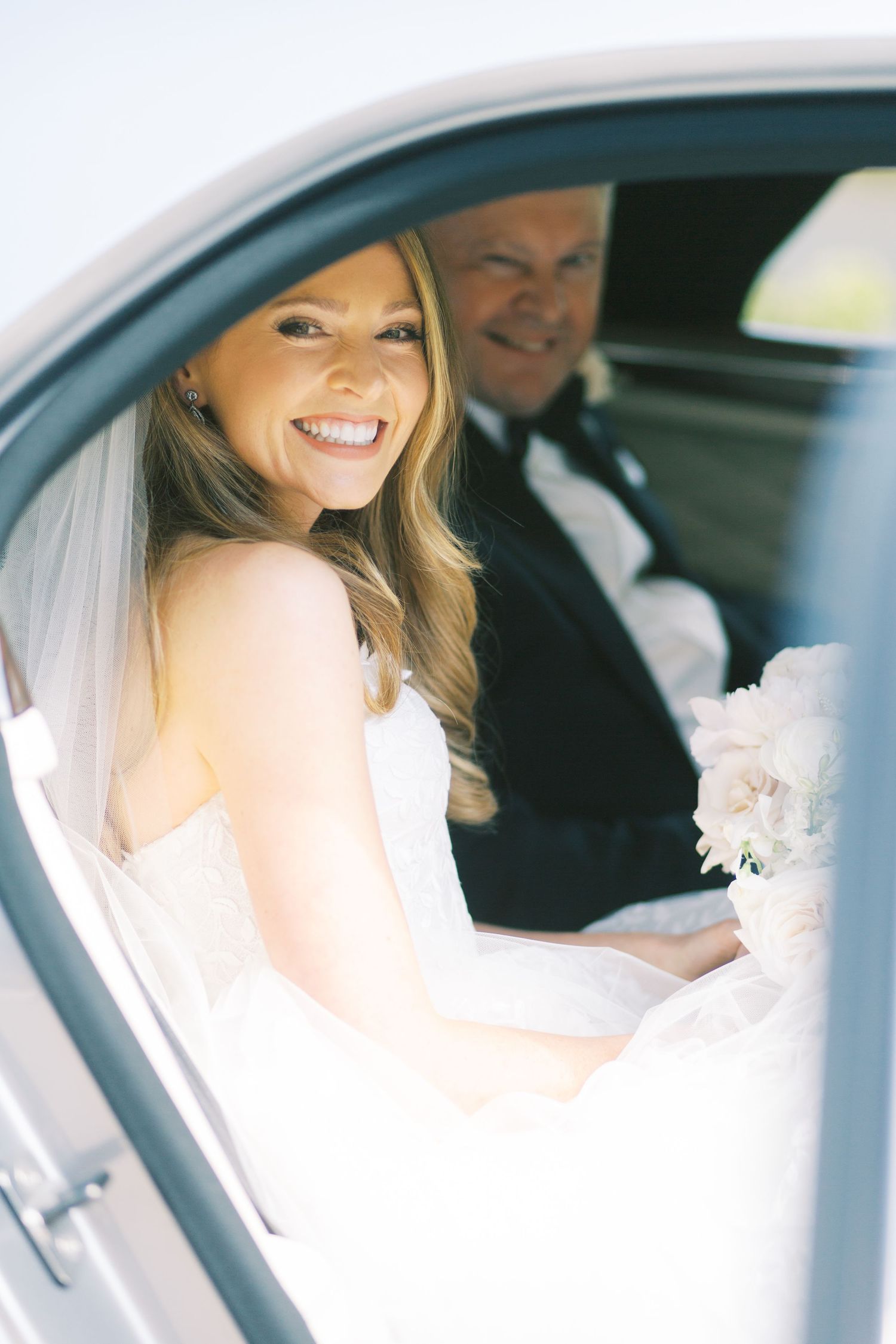 Smiling bride in white gown and veil sits in car holding bouquet with groom in tuxedo behind her.