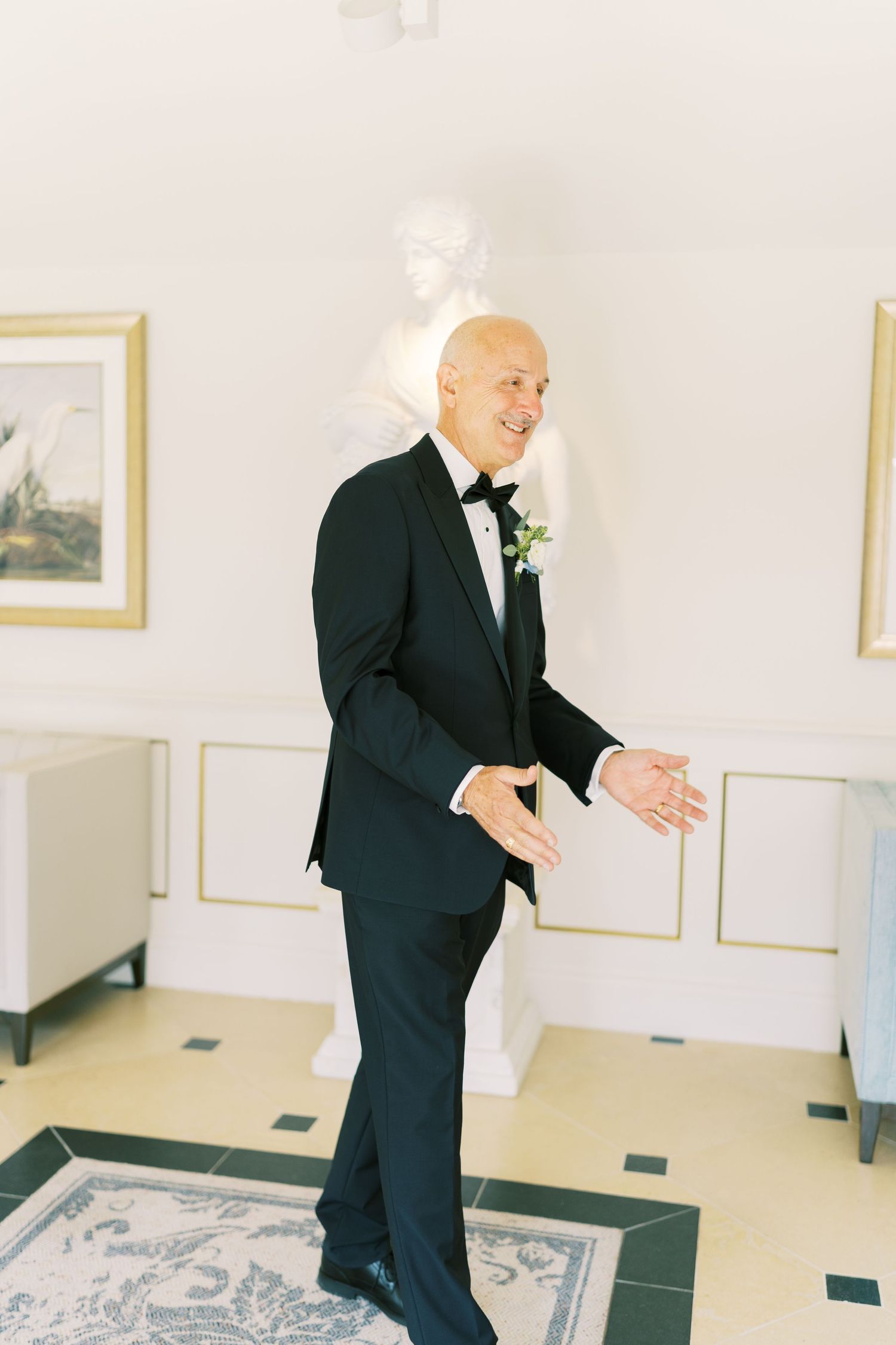 Smiling groom in black tuxedo with boutonniere clapping hands in elegant white room with classical statue.