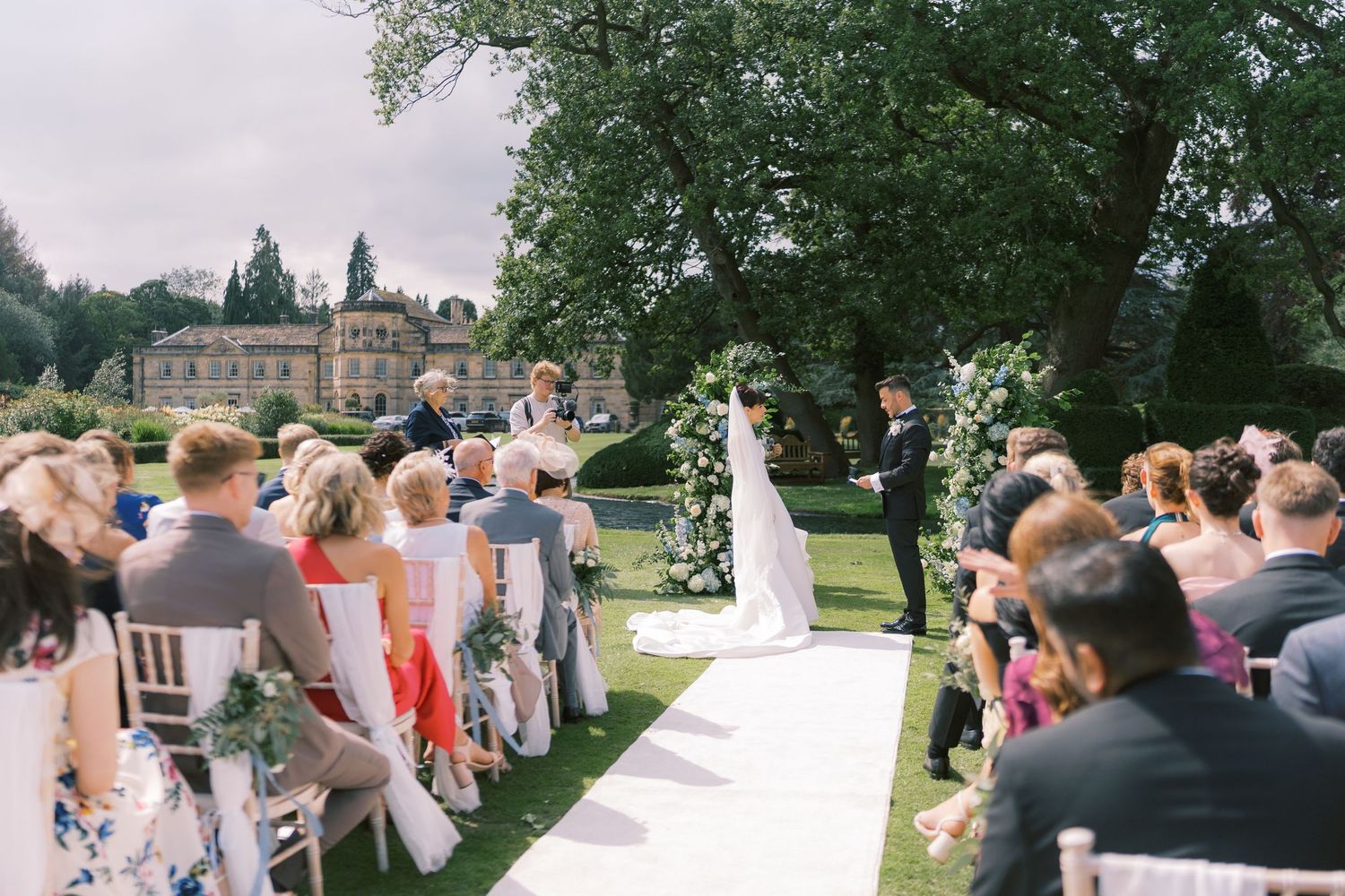 Outdoor wedding ceremony on white aisle in lush garden with guests seated, officiant and couple at floral arch, historic manor behind.