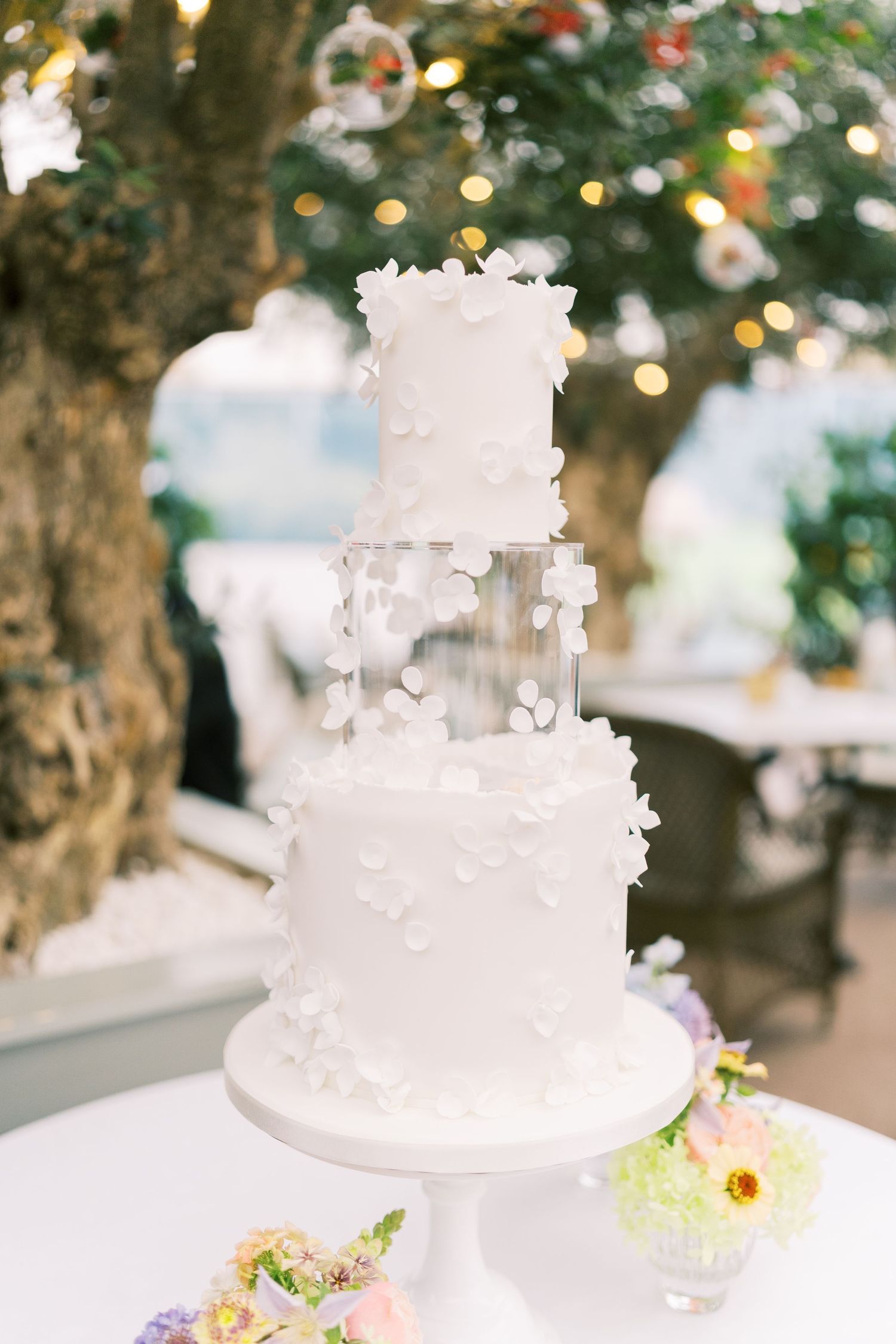 Elegant three-tier white wedding cake adorned with delicate floral appliques on a cake stand outdoors.