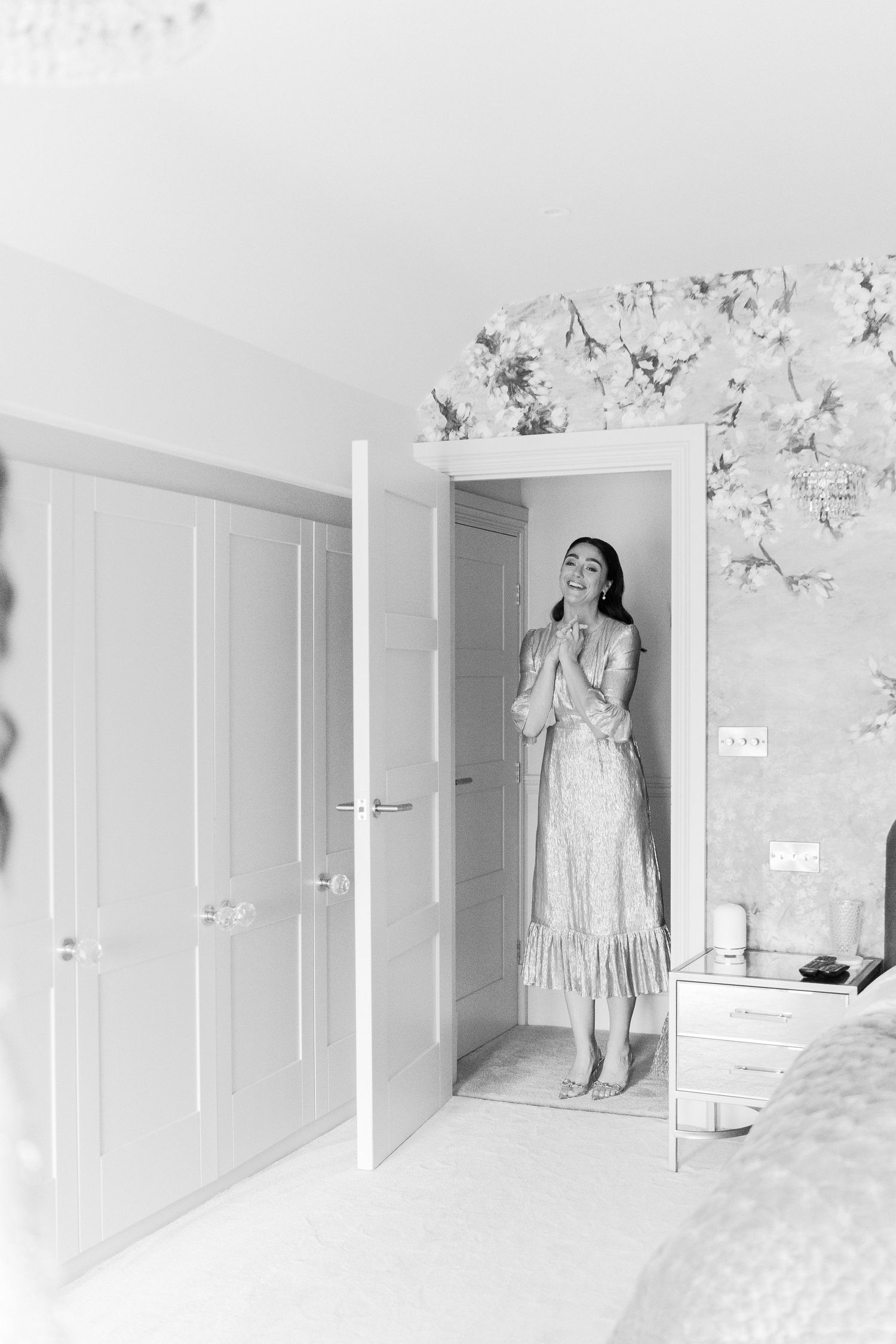 Black and white photo of a bride in a lace dress standing in a doorway with floral wallpaper.