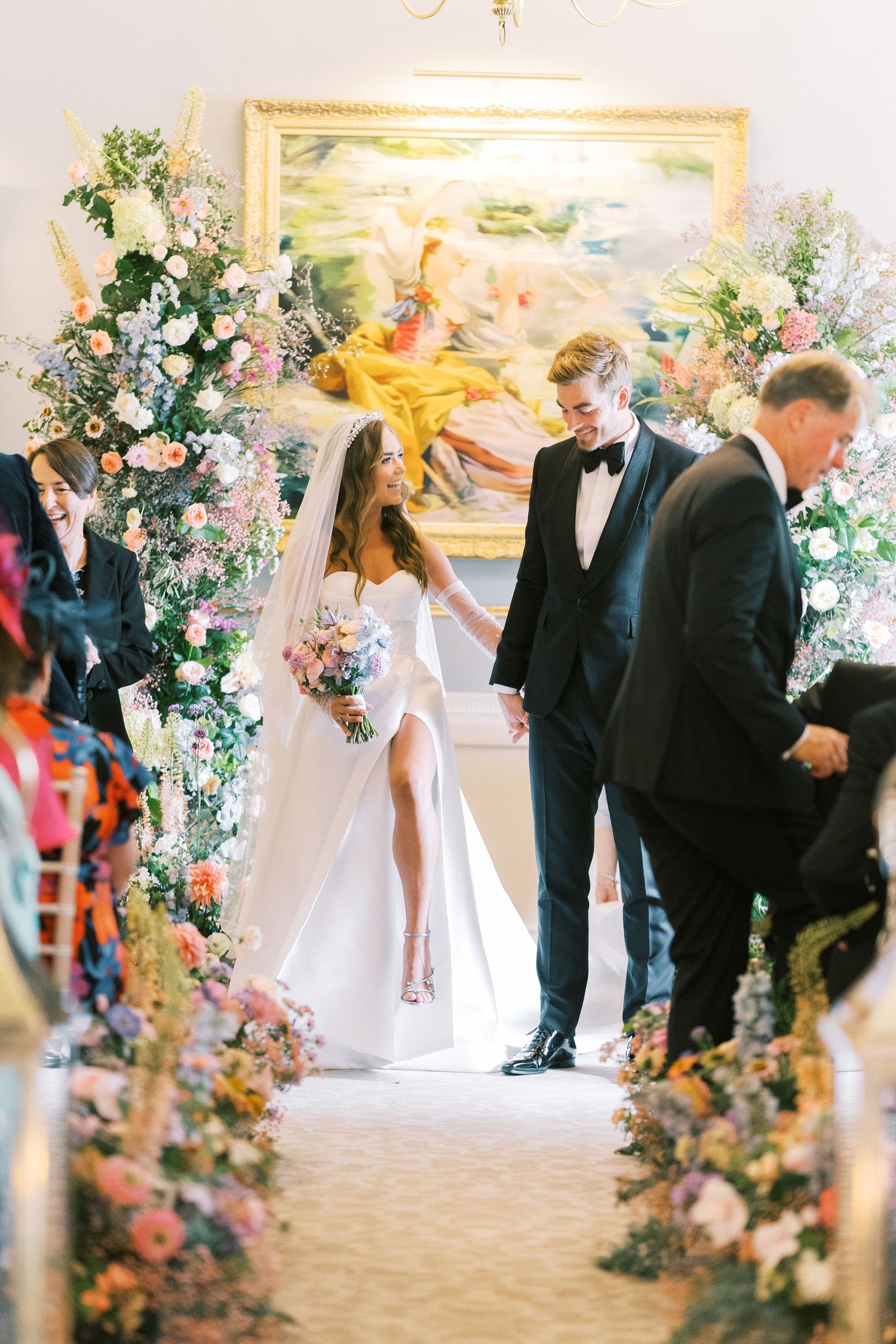 Newlyweds walking down aisle lined with colorful floral arrangements after wedding ceremony in decorated hall.