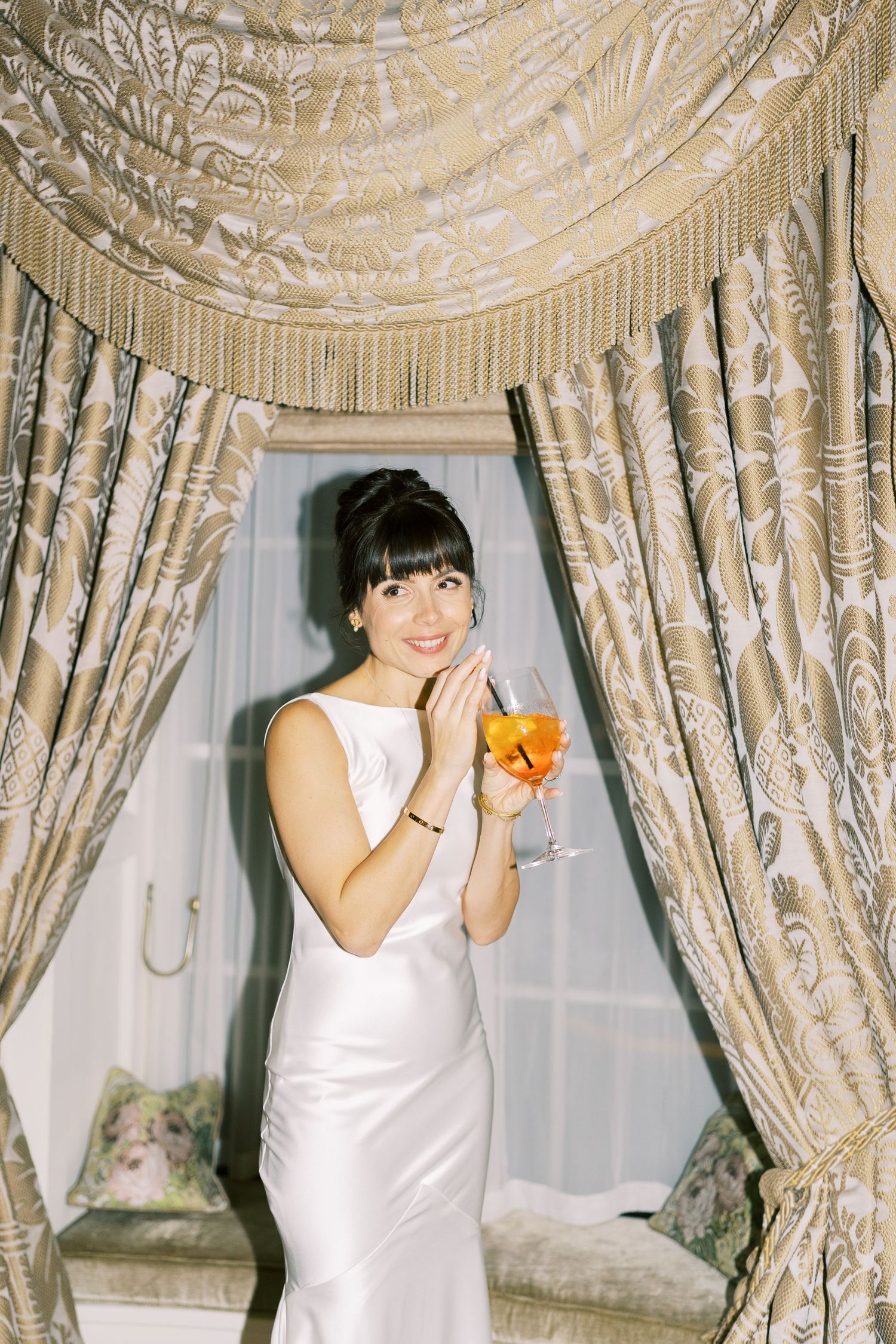 Elegant bride in white satin gown holding orange cocktail, smiling before ornate gold damask curtains.