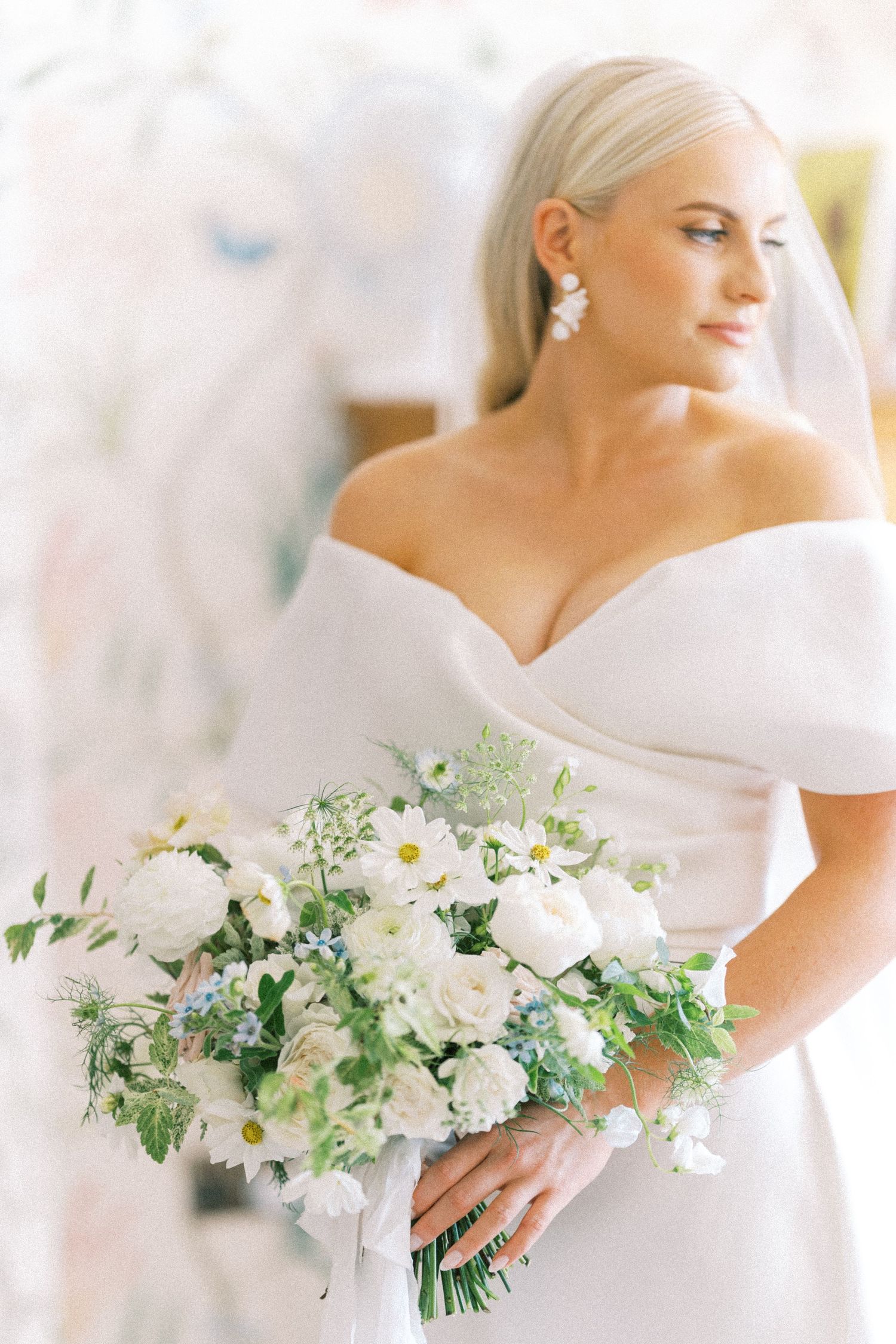 Elegant blonde bride in off-shoulder white gown holding a lush white and green wedding bouquet.