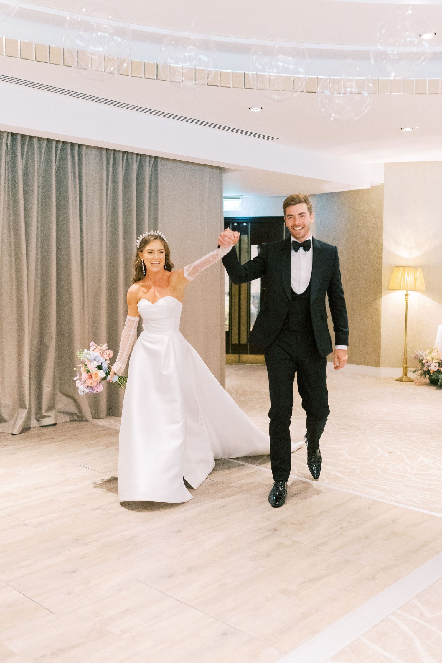 Joyful newlyweds make grand entrance at wedding reception, bride in white gown holding bouquet, groom in black tuxedo.