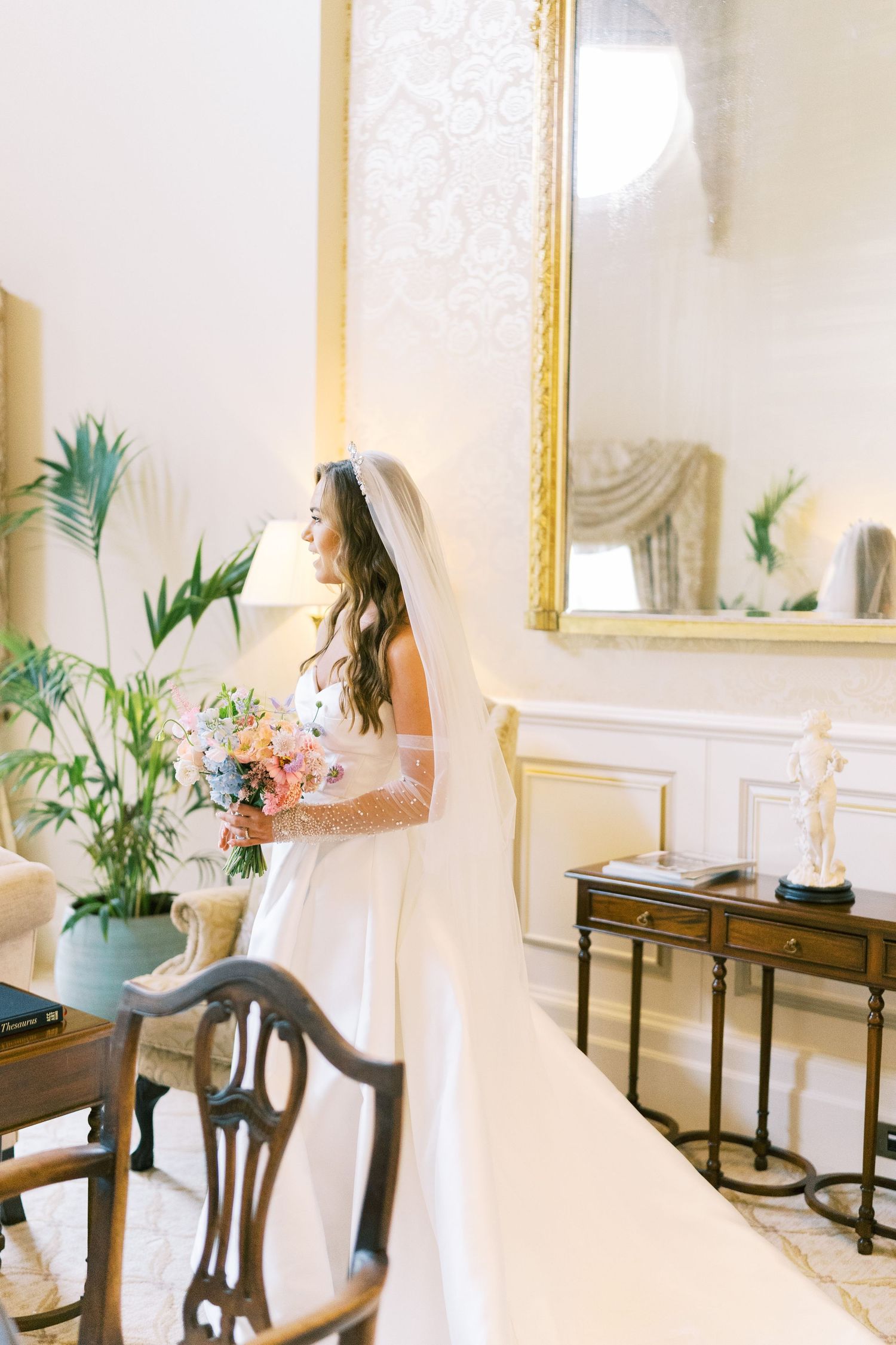 Bride in white gown and veil holds colorful bouquet in elegant room with gold mirror and green plants.