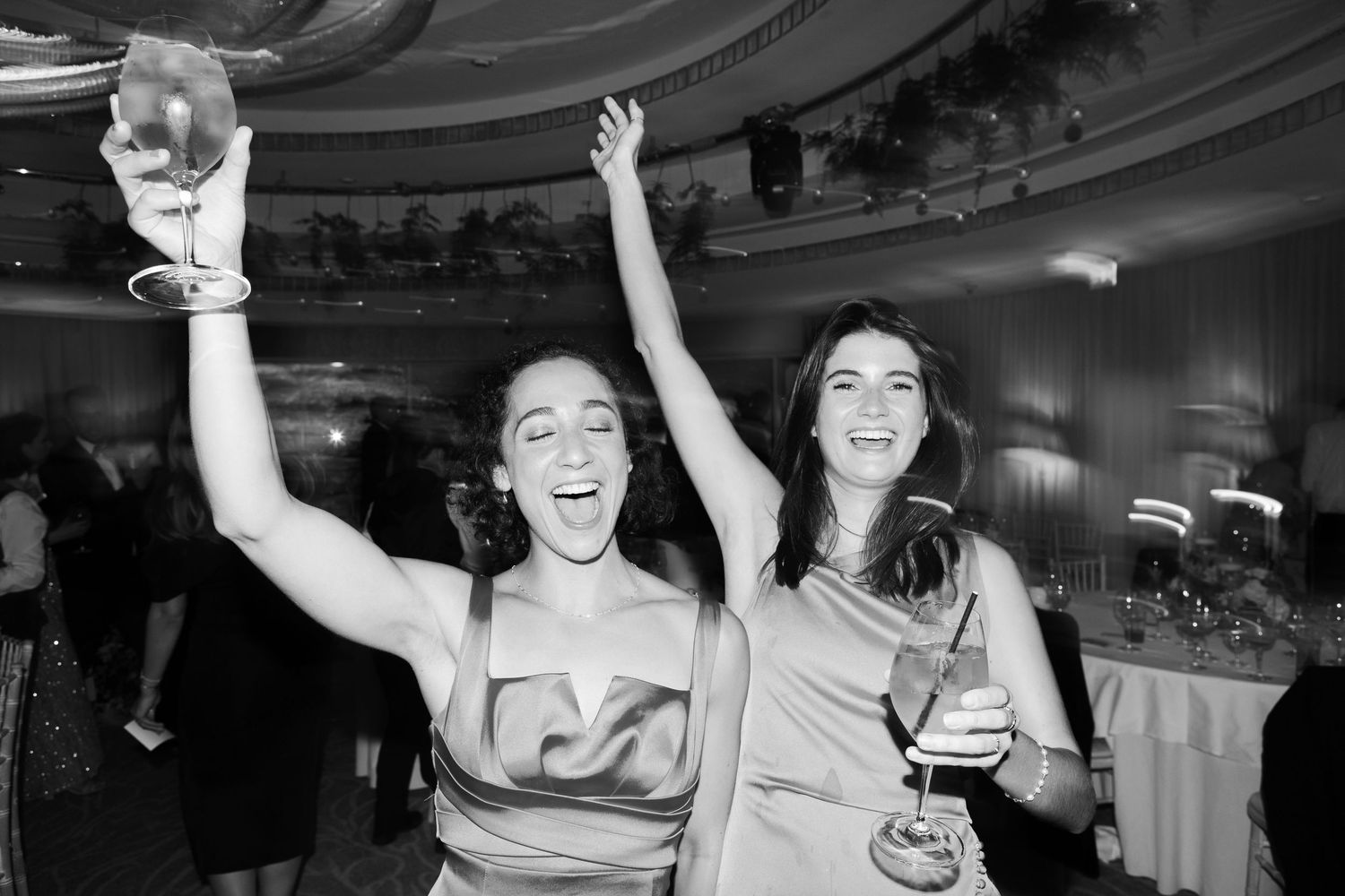 Two women in elegant gowns celebrate joyfully with drinks at a formal black and white event.