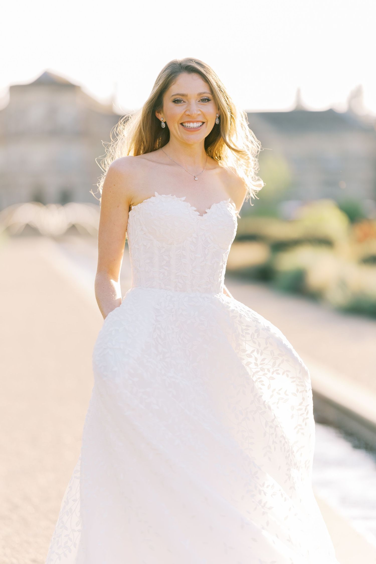 Smiling bride in a strapless white lace ball gown posing outdoors in golden sunlight.