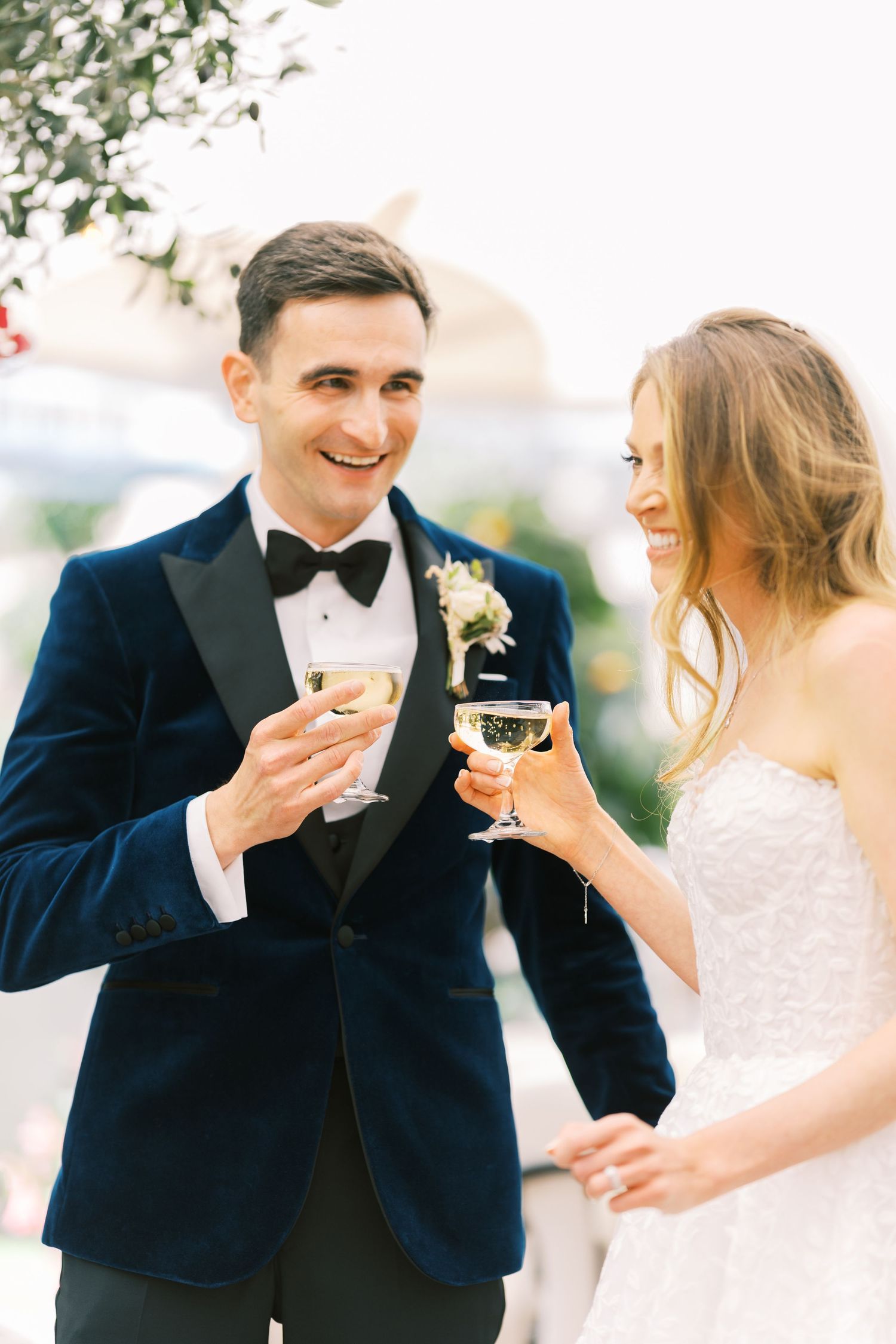 Smiling bride and groom in navy tuxedo clinking champagne glasses at wedding reception.