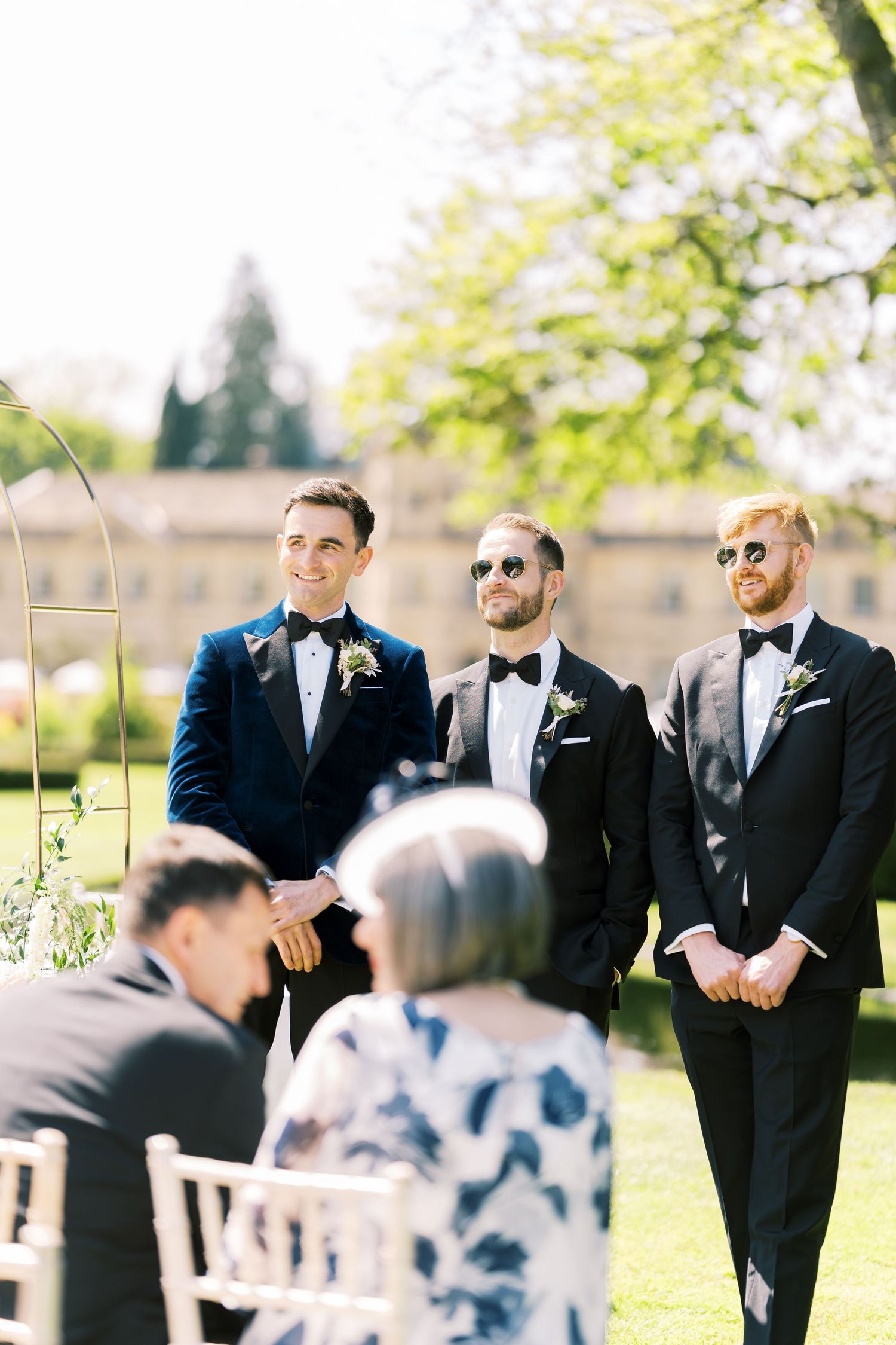 Groom in navy tuxedo smiles at altar flanked by groomsmen in black tuxedos at outdoor wedding ceremony.