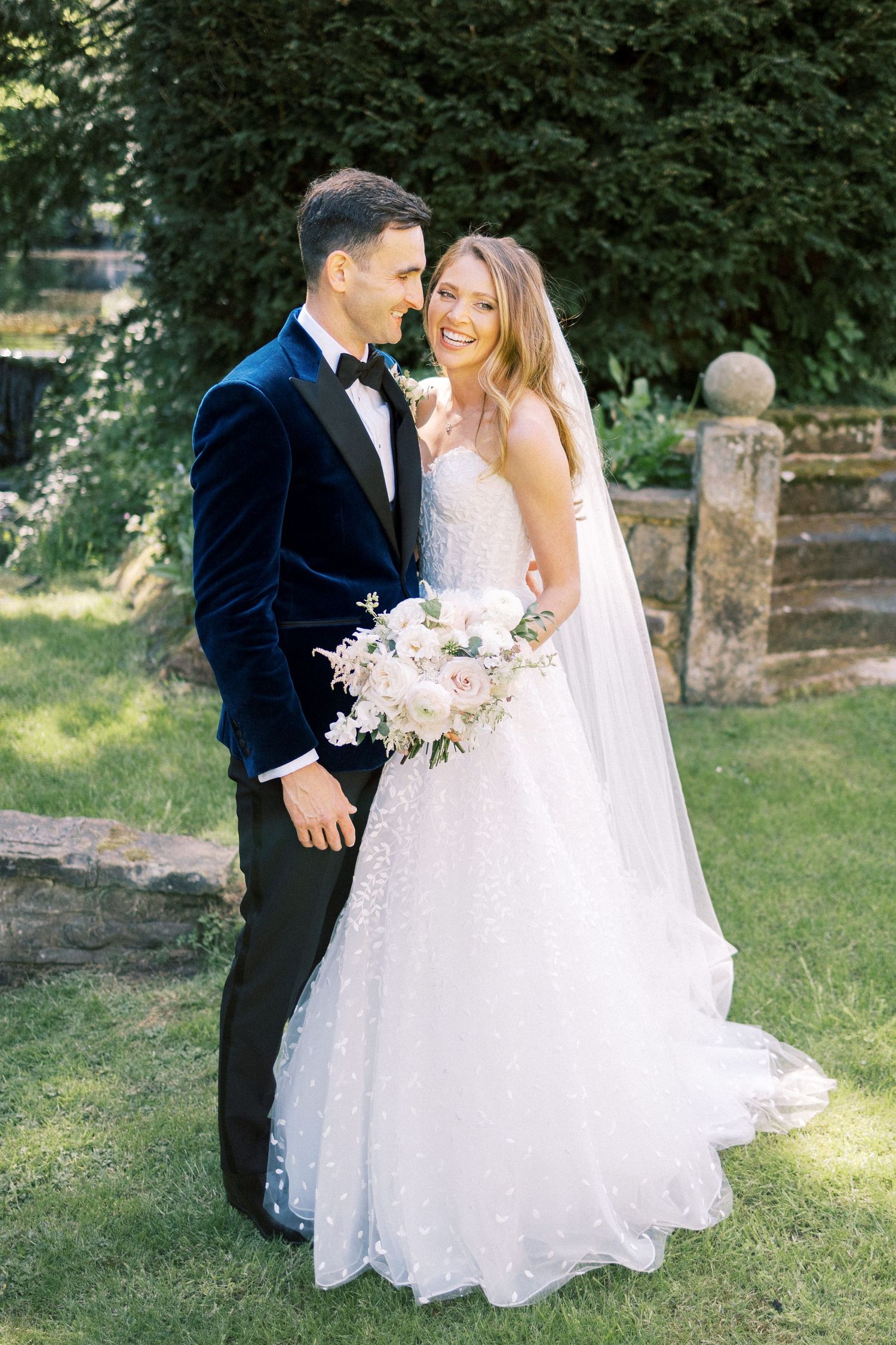 Bride and groom pose outdoors in garden, she holds blush bouquet in beaded gown, he wears navy velvet tuxedo jacket.