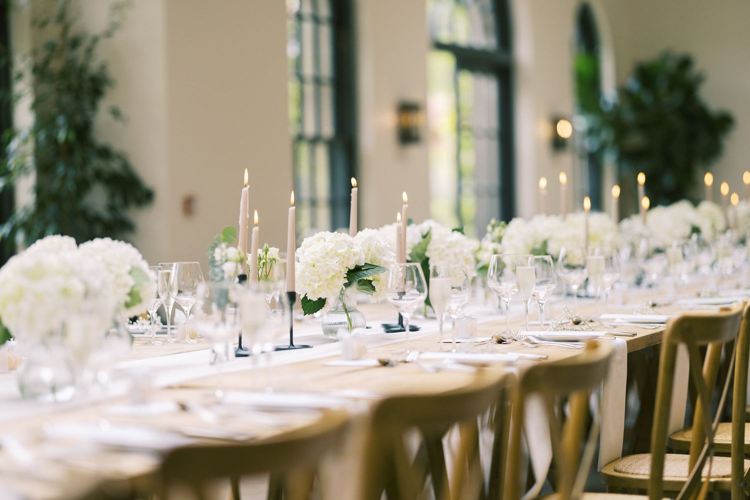 Elegant wedding reception table with white hydrangea centerpieces, tall taper candles, and crystal glassware in a bright venue.