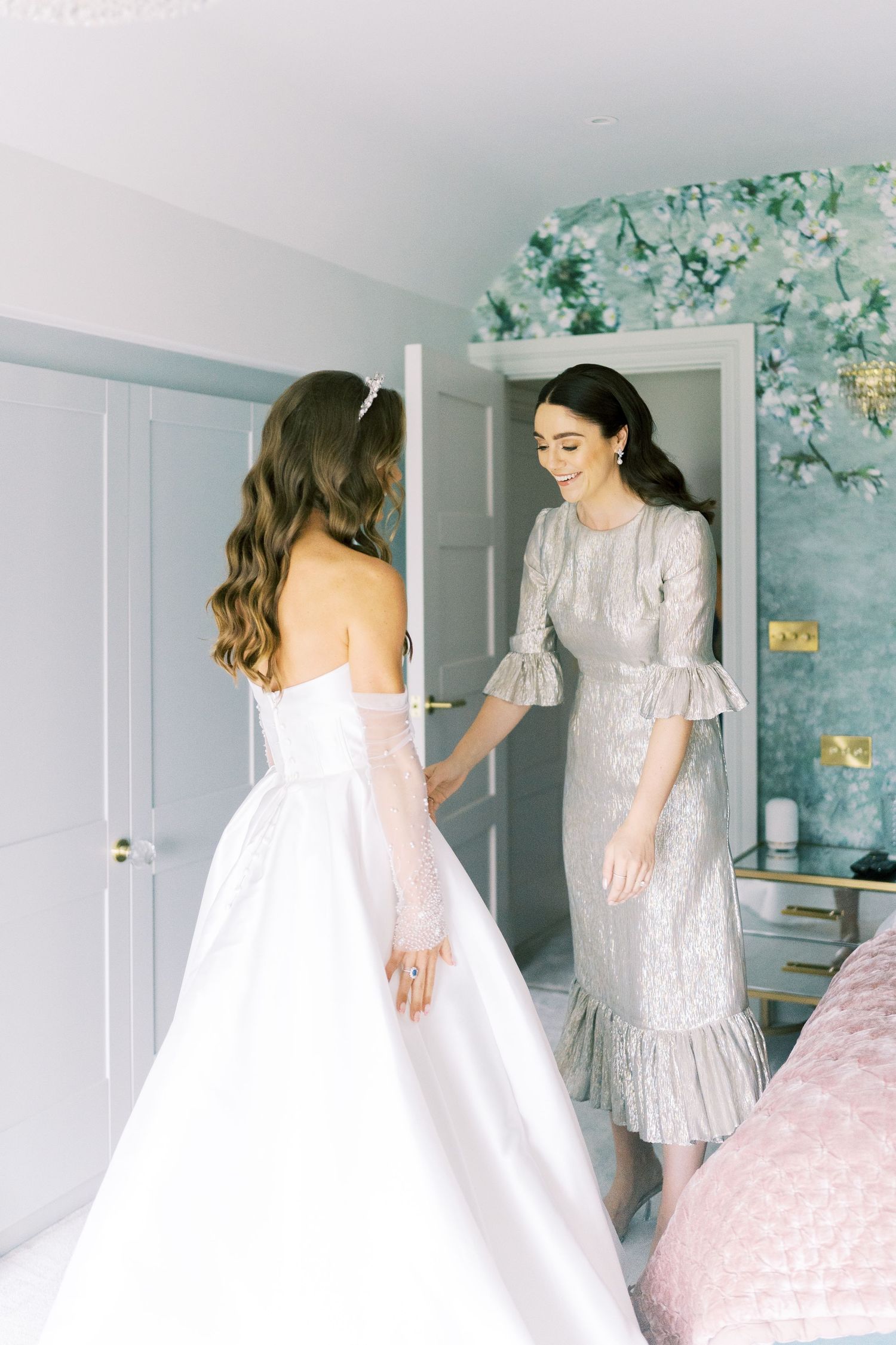 Bride in off-shoulder white wedding gown with sheer sleeves holds hands with smiling bridesmaid in silver midi dress.
