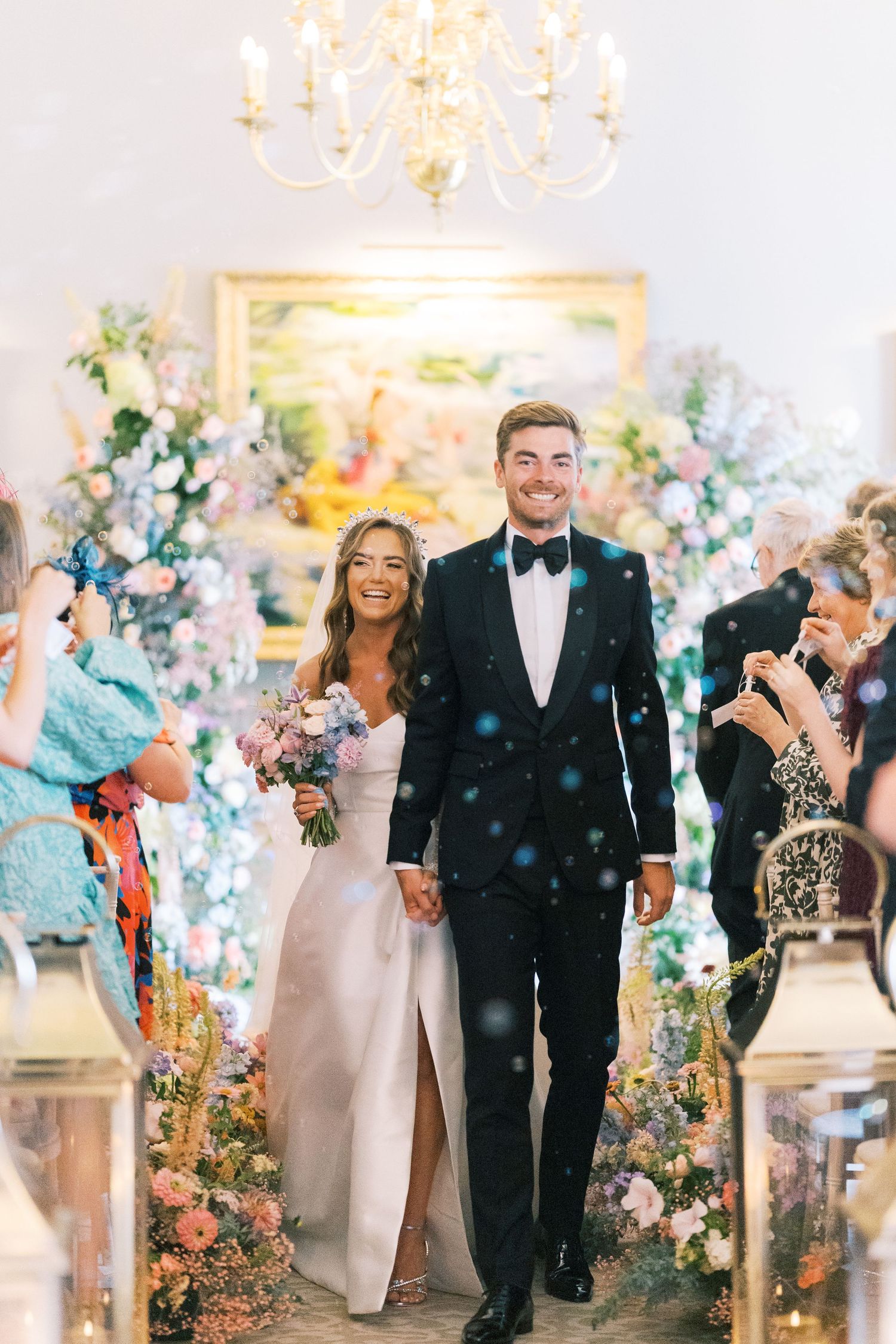 Bride and groom walk down aisle after wedding ceremony, surrounded by floral arrangements and confetti in elegant venue.