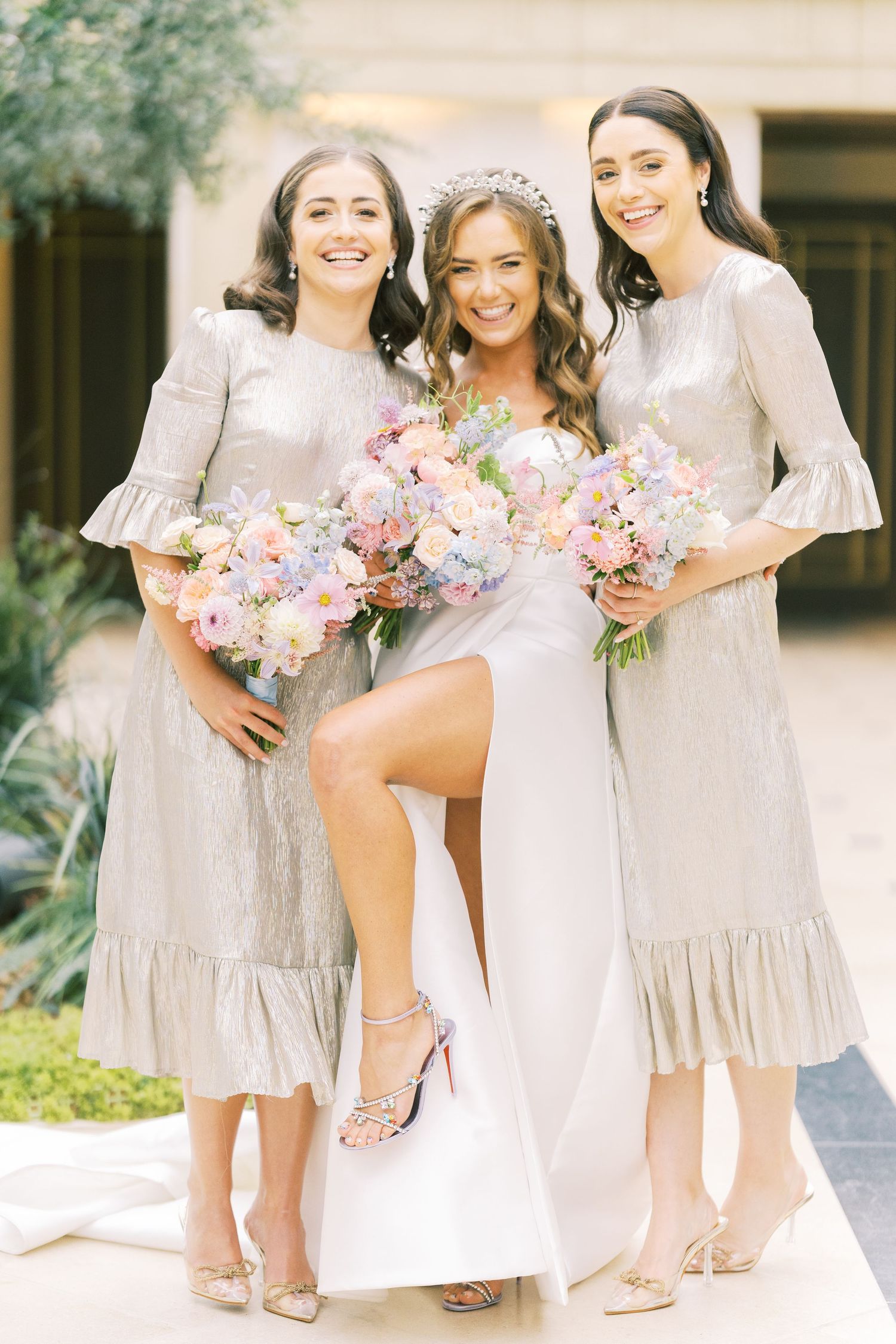 Bride in white gown with tiara and two bridesmaids in silver dresses holding pastel bouquets outdoors.