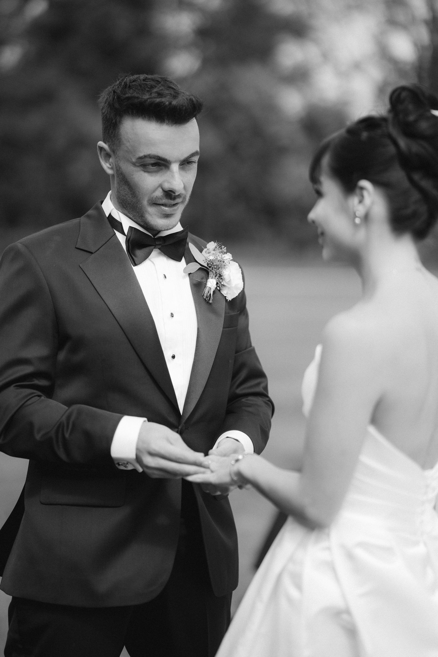 Groom placing wedding ring on bride's finger during outdoor ceremony in black and white photo.