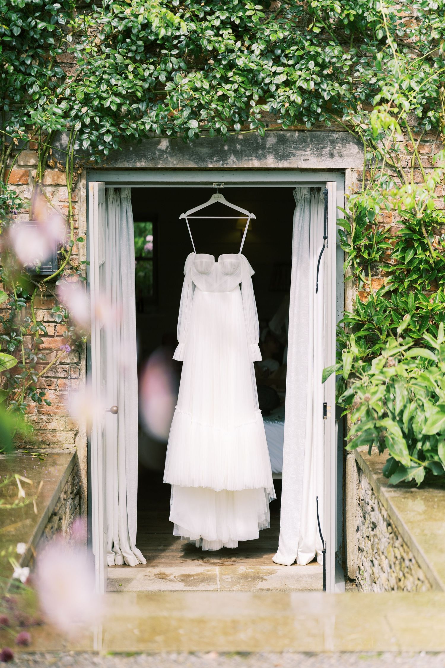 White wedding dress hanging in a doorway surrounded by lush green ivy and pink flowers at an elegant venue.