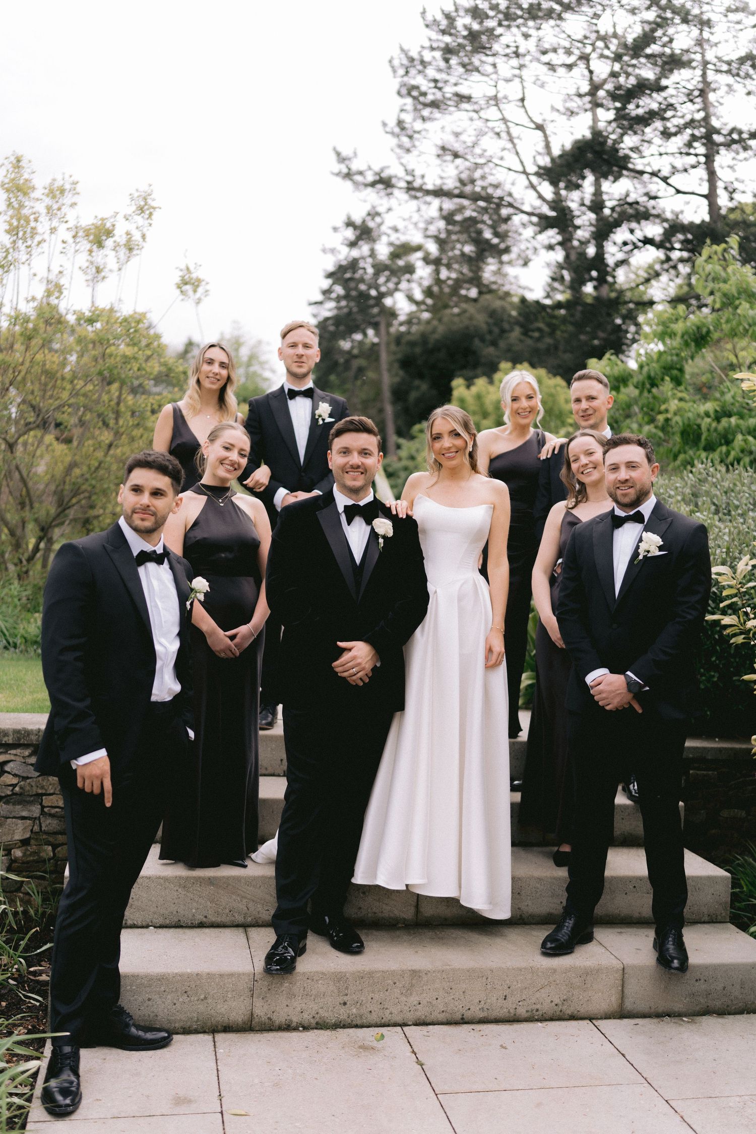 Wedding party poses on stone steps, bride in white gown with groom and bridal party in black formal attire outdoors.