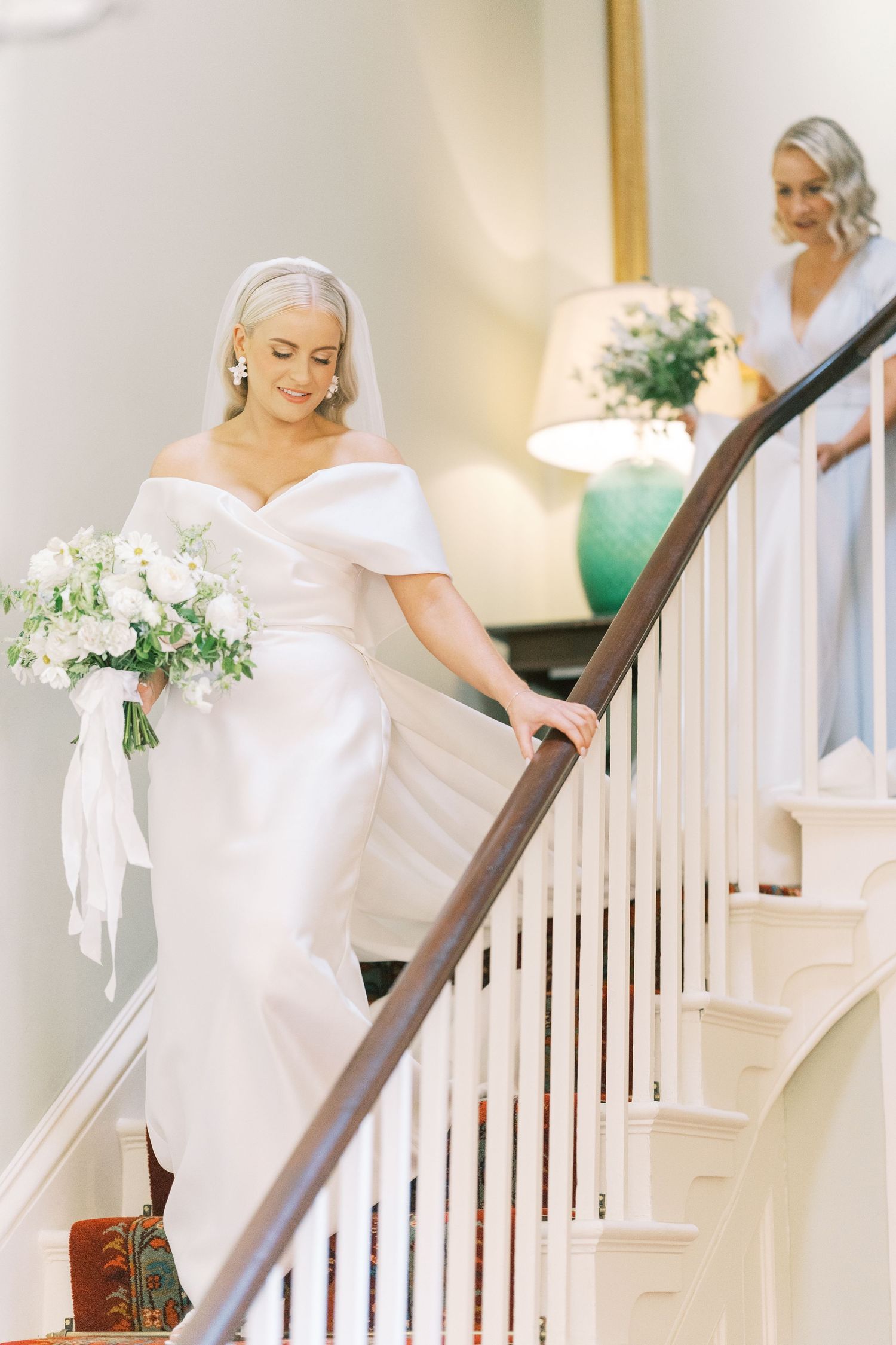 Bride in off-shoulder white gown descending elegant staircase while holding white floral bouquet.