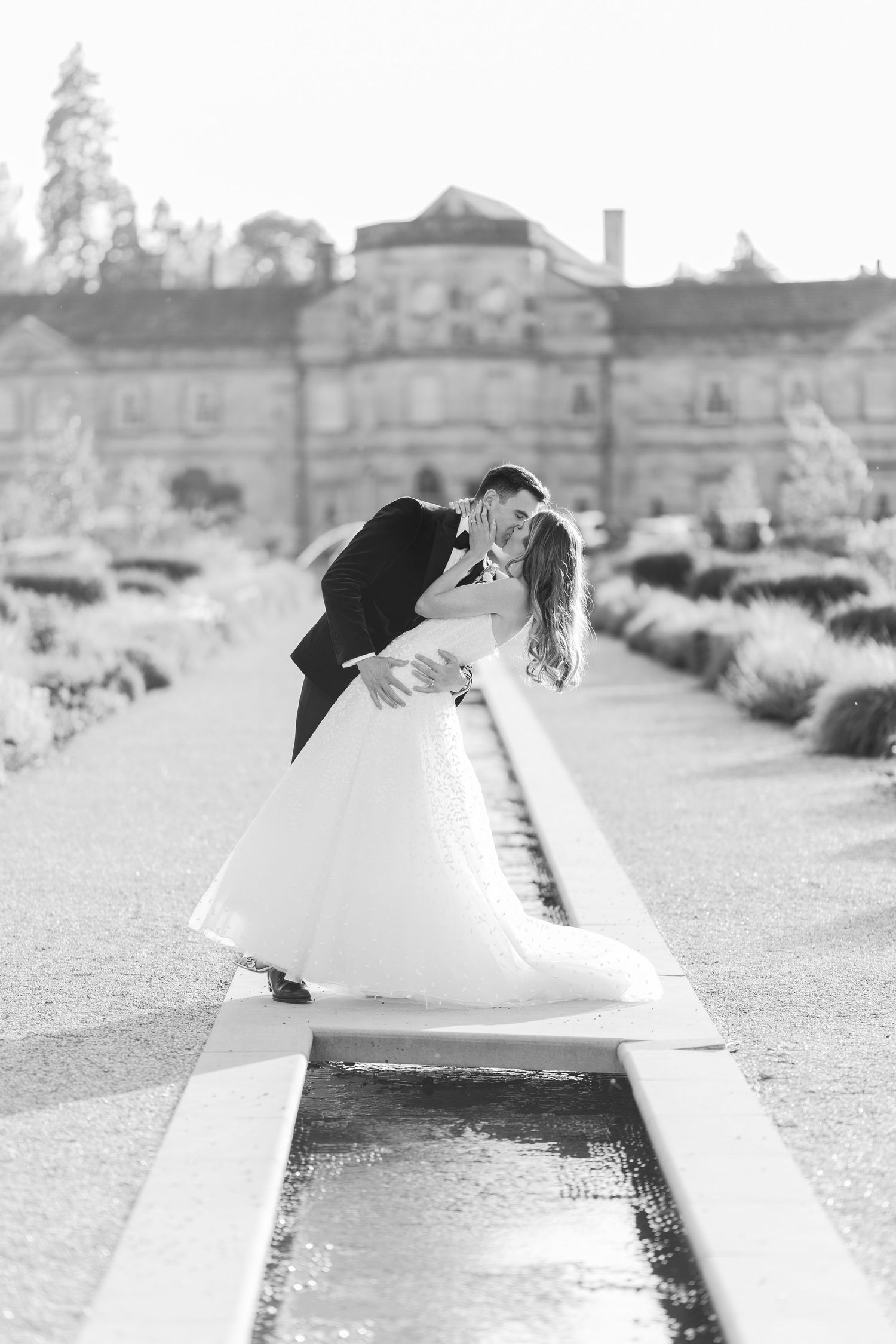 Black and white photo of bride and groom sharing a romantic dip kiss beside a garden fountain at an elegant estate.