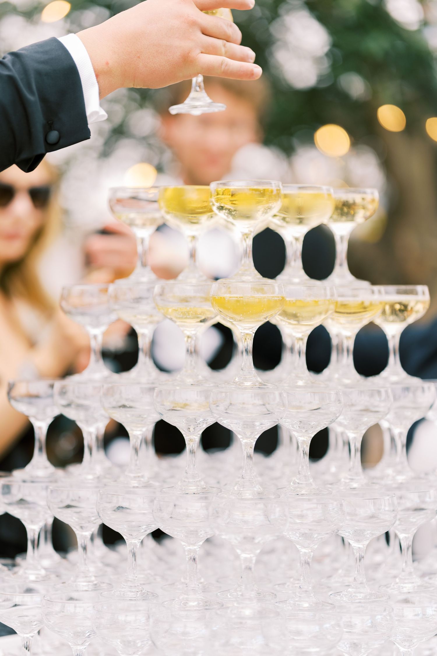 Hand reaching for a champagne coupe from a sparkling tiered champagne tower at a wedding reception.